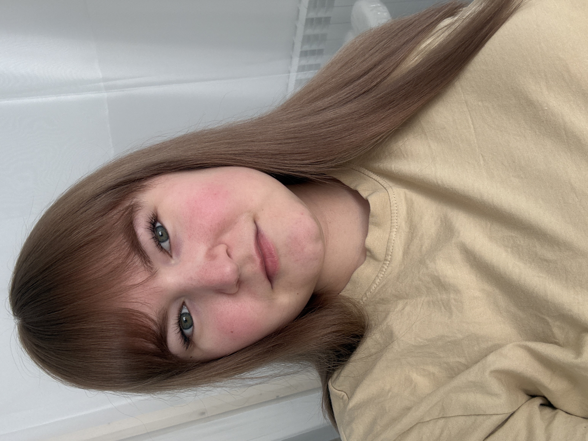 A young woman with light skin, light brown hair with bangs, and green eyes is pictured indoors. Her cheeks appear flushed. She is wearing a light tan t-shirt. The background is blurred, showing a white wall and a possible window with blinds. There is no verifiable evidence to pinpoint the location as Chaikovsky, Russia. The primary subject is the woman and her immediate surroundings.