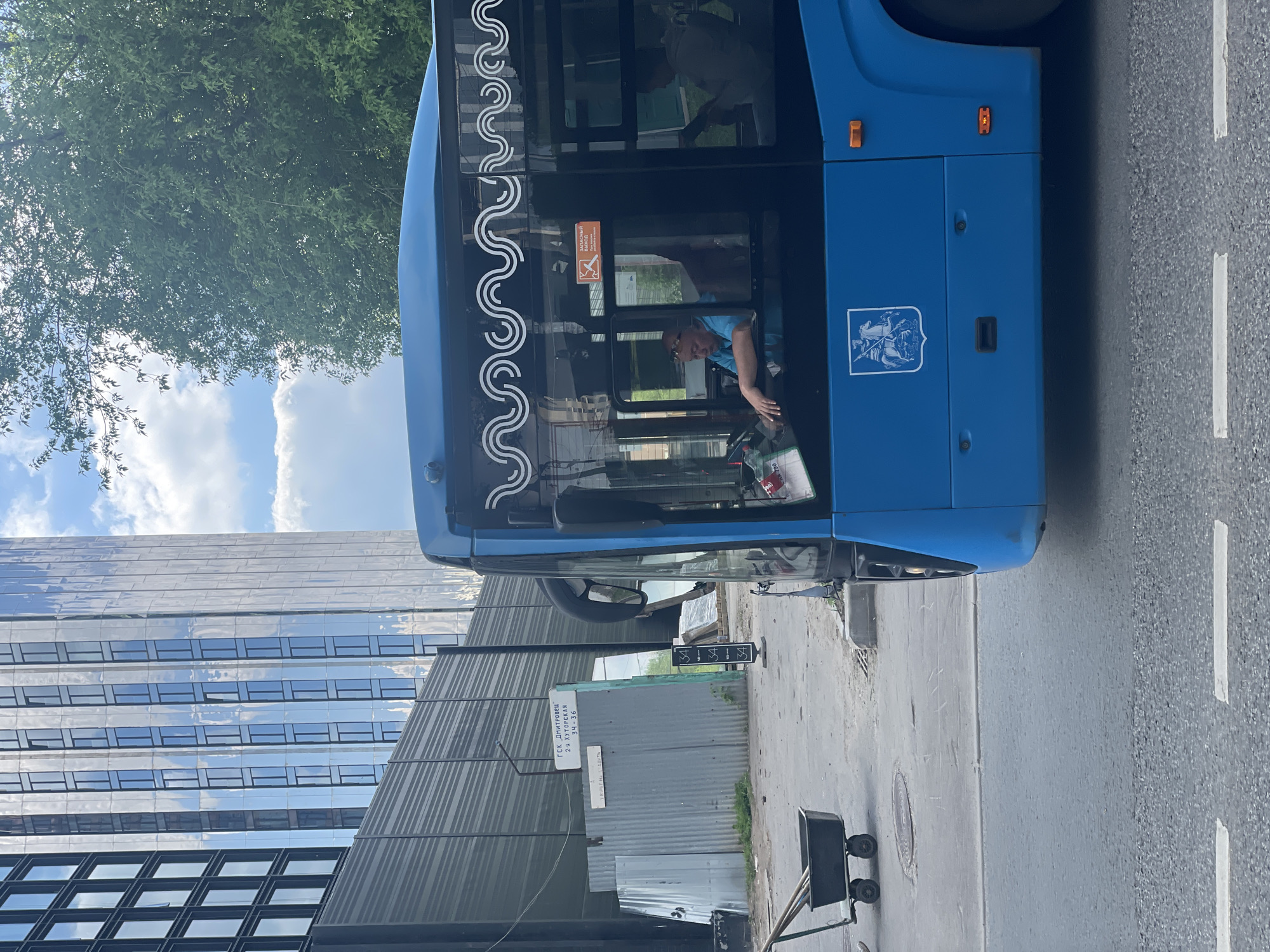 The image shows a blue bus on a street in Khimki, Russia. A man in a blue shirt is in the driver's seat. In the background are a building with a reflective glass facade, trees, and cloudy skies. A metal fence and signage with Cyrillic text are visible. A black wheeled cart sits on the sidewalk. The street has white lines indicating lanes. The bus has a white emblem and decorative designs. The bus has a sign indicating the emergency exit.