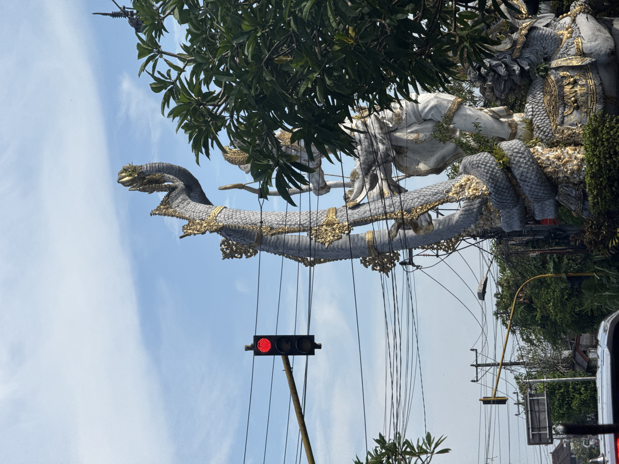 The image displays a grand, ornate sculptural monument under a clear blue sky, likely situated in the Ubud District of Indonesia. The central feature is a massive, multi-headed serpent-like creature, possibly a Naga, which is predominantly gray and white with intricate gold detailing and embellishments. Its serpentine body forms a large, curving structure, suggesting it is part of a grand gate or archway.
Perched upon and integrated into the serpent are large, imposing figures, also in white and gray with gold accents. These figures appear to be deities or mythological heroes, with one seemingly holding a staff or spear and another exhibiting textured, scaled armor. Lush green foliage from surrounding trees frames the monument, with branches and leaves partially obscuring some parts of the sculpture and sky.
In the lower left portion of the image, a red traffic light is prominently visible, indicating a stop. It is mounted on a brown pole, and numerous dark utility wires crisscross the sky, extending from various points above the street. A small section of a vehicle, possibly a car, is visible on the far right edge of the frame. The scene is brightly illuminated by natural light, indicating a clear and sunny daytime, with only a few scattered white clouds visible in the expanse of blue sky. No explicit activity is occurring, and no text is visible.