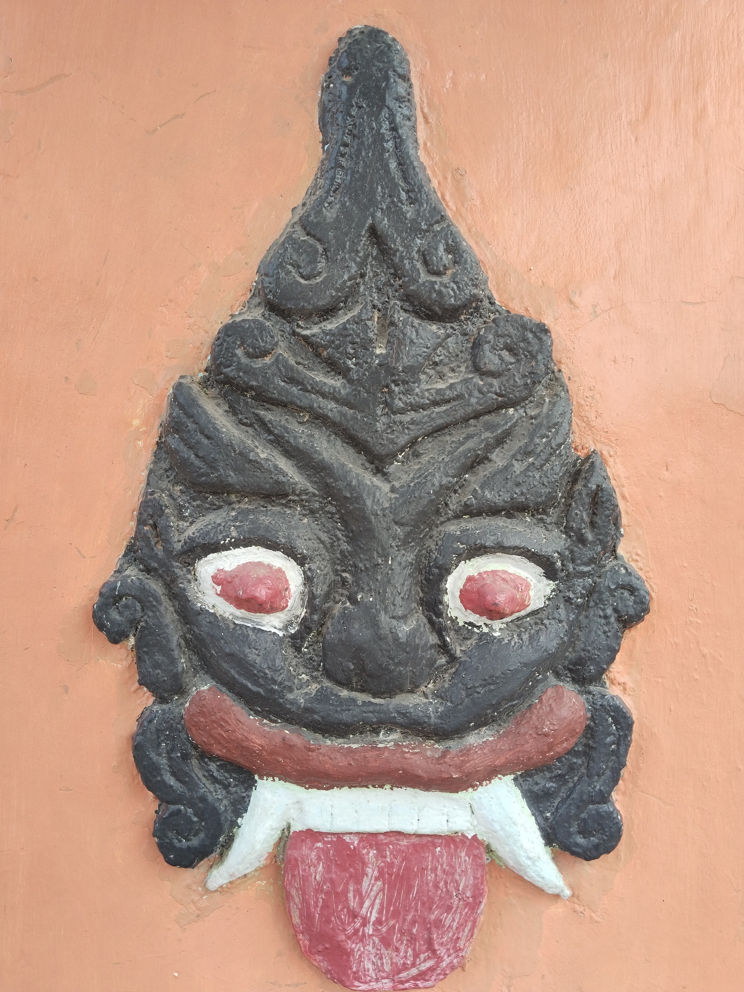 A carved, painted mask is mounted on a peachy-orange wall in Sidomukti, Indonesia. The mask is vertically oriented, with a black face, red and white eyes, and a red mouth with two protruding white teeth. A red tongue sticks out from the mouth. The top of the mask has a carved design that tapers to a point. The background wall is a solid color, with slight surface variations.