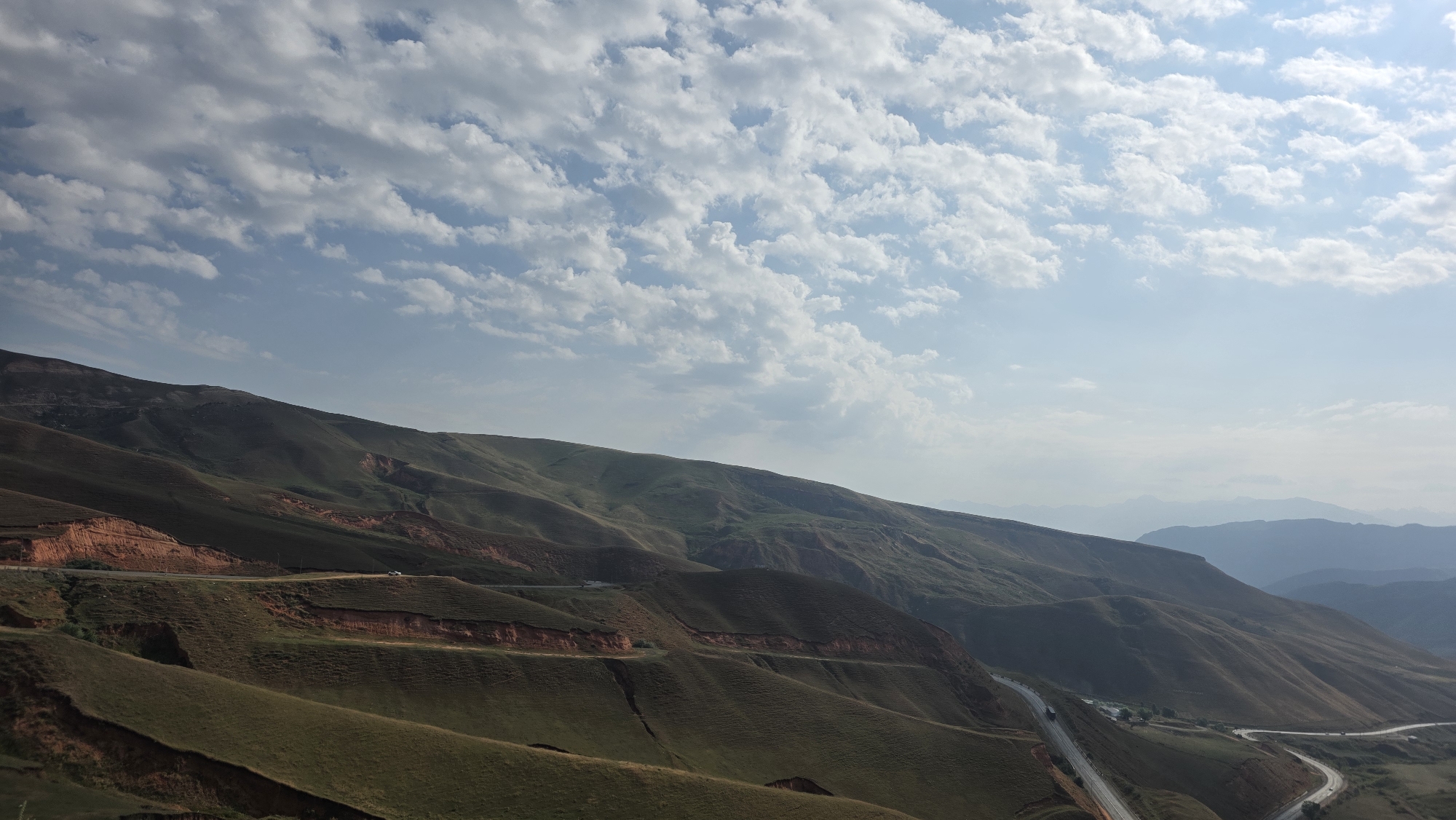 The image shows a vast, rolling mountain landscape under a partly cloudy sky. The mountains are covered in a mixture of green grass and reddish-brown soil, with visible erosion patterns on some slopes. A winding road is carved into the mountainside, with a few vehicles visible on it. The scene appears to be in a rural, mountainous region. The specific location is Taldyk, Kyrgyzstan.