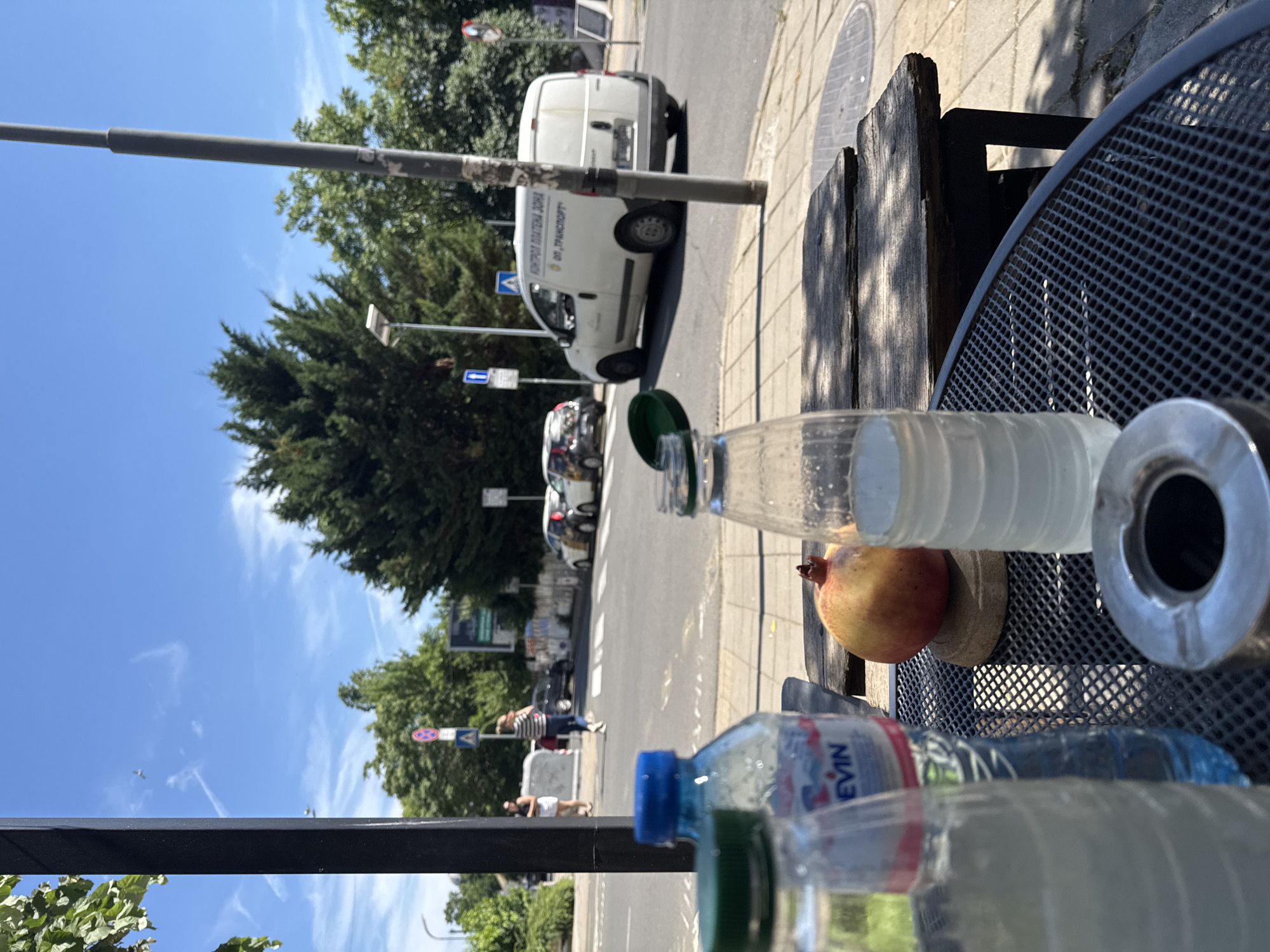 The image captures a sunny outdoor scene in Burgas, Bulgaria, likely near a cafe or restaurant by a street. In the foreground, on a dark metal table, are several items: an ashtray, two partially filled plastic water bottles (one with a blue cap and Evian branding, the other with a green cap), and a pomegranate resting on a small wooden coaster. Behind the table, a wooden bench is visible.
The middle ground shows a street with a pedestrian crossing. Several cars are on the road, including a white van with Cyrillic text on its side that appears to read "КОНТРОЛ ПЛАТЕНА ЗОНА" and "ОП. ТРАНСПОРТ". Two people are on the sidewalk, one walking and the other standing. Traffic signs and streetlights are also present.
The background is dominated by lush green trees under a bright blue sky with scattered white clouds. The overall atmosphere is casual and relaxed, suggesting a moment of repose on a warm day in Burgas.