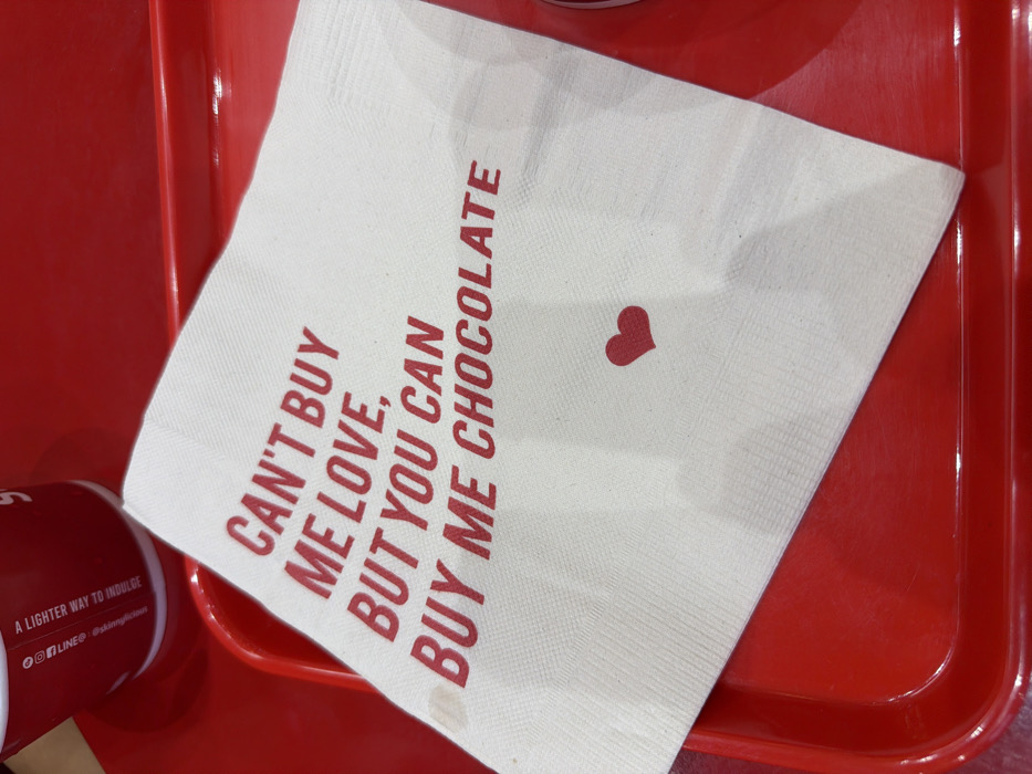 A white paper napkin, featuring the red text "CAN'T BUY ME LOVE, BUT YOU CAN BUY ME CHOCOLATE" and a small red heart, rests on a glossy red plastic tray. A red paper cup is partially visible in the upper left corner of the frame, bearing the text "A LIGHTER WAY TO INDULGE" and the social media handle "@slidingprawn." This indicates the setting is a Sliding Prawn restaurant, located in the city of Bangkok, Thailand. The napkin exhibits a minor discolored smudge on its lower left side. The scene is captured under uniform artificial lighting, with no individuals or active interactions present.