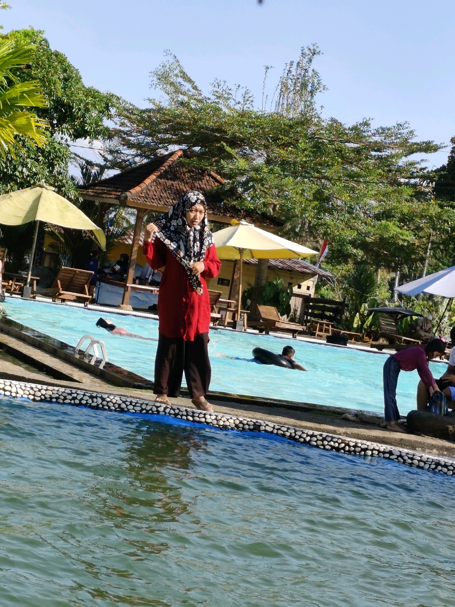 This is a full, eye-level shot of a swimming pool area on a sunny day. In the foreground, the water is green and rippling. The pool's edge is decorated with a border of white and gray pebbles. A woman wearing a red tunic and dark pants, with a patterned headscarf, stands at the pool's edge, adjusting her scarf. In the pool, one person is swimming with a black inflatable ring. Further back, several lounge chairs and umbrellas are visible, as well as a small building with a brown tiled roof. Lush green trees and vegetation surround the area. The sky is clear and blue. The location is likely a public pool or resort in a tropical or subtropical region of Indonesia.