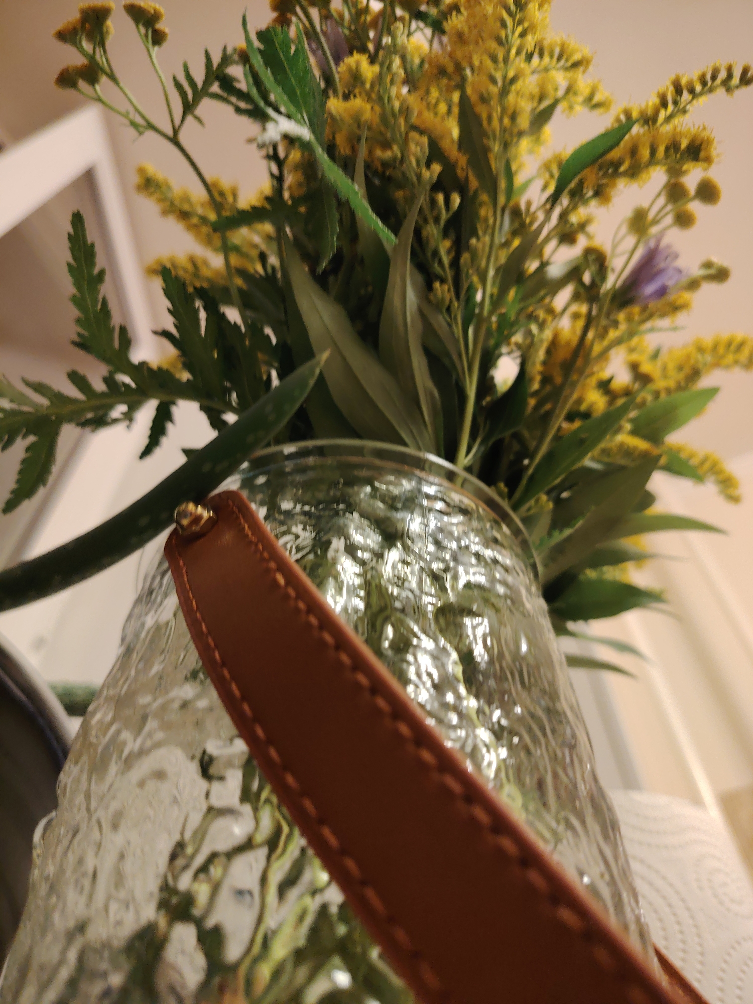 The image shows a close-up, low-angle shot of a bouquet of yellow flowers with green foliage in a textured glass vase. A brown leather strap with stitching is visible across the front of the vase, secured by a gold stud. The background is softly blurred, showing a light-colored wall or surface and what appears to be a roll of paper towels. The scene is indoors, likely in a residential setting. The city of Konakovo, Russia, is not depicted or suggested in the image.