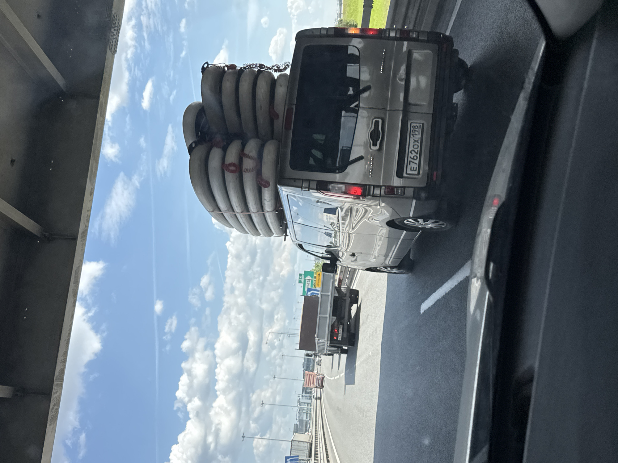 This vertically oriented image captures a bright daytime scene on a multi-lane highway, viewed from inside a moving vehicle. The most prominent subject is a silver van traveling in an adjacent lane, heavily laden with numerous light-colored, cylindrical objects, likely pipes or conduits, stacked high on its roof rack and secured with red and black straps.
The rear of the van is clearly visible, including its Russian license plate, "E762OX 198," which indicates registration in the Saint Petersburg/Leningrad Oblast region. The van's rear windows are tinted, and its taillights are illuminated. Further down the highway, a smaller flatbed truck can be seen.
The sky overhead is bright blue with scattered white, puffy clouds, suggesting clear and sunny weather. To the far left, the concrete underside of an overpass or bridge structure is partially visible. The highway itself is well-maintained with clear white lane markings. In the distance, a green landscape borders the road, and road signs with Cyrillic text can be faintly discerned, confirming the location as Russia. The overall scene depicts highway traffic under good weather conditions.
