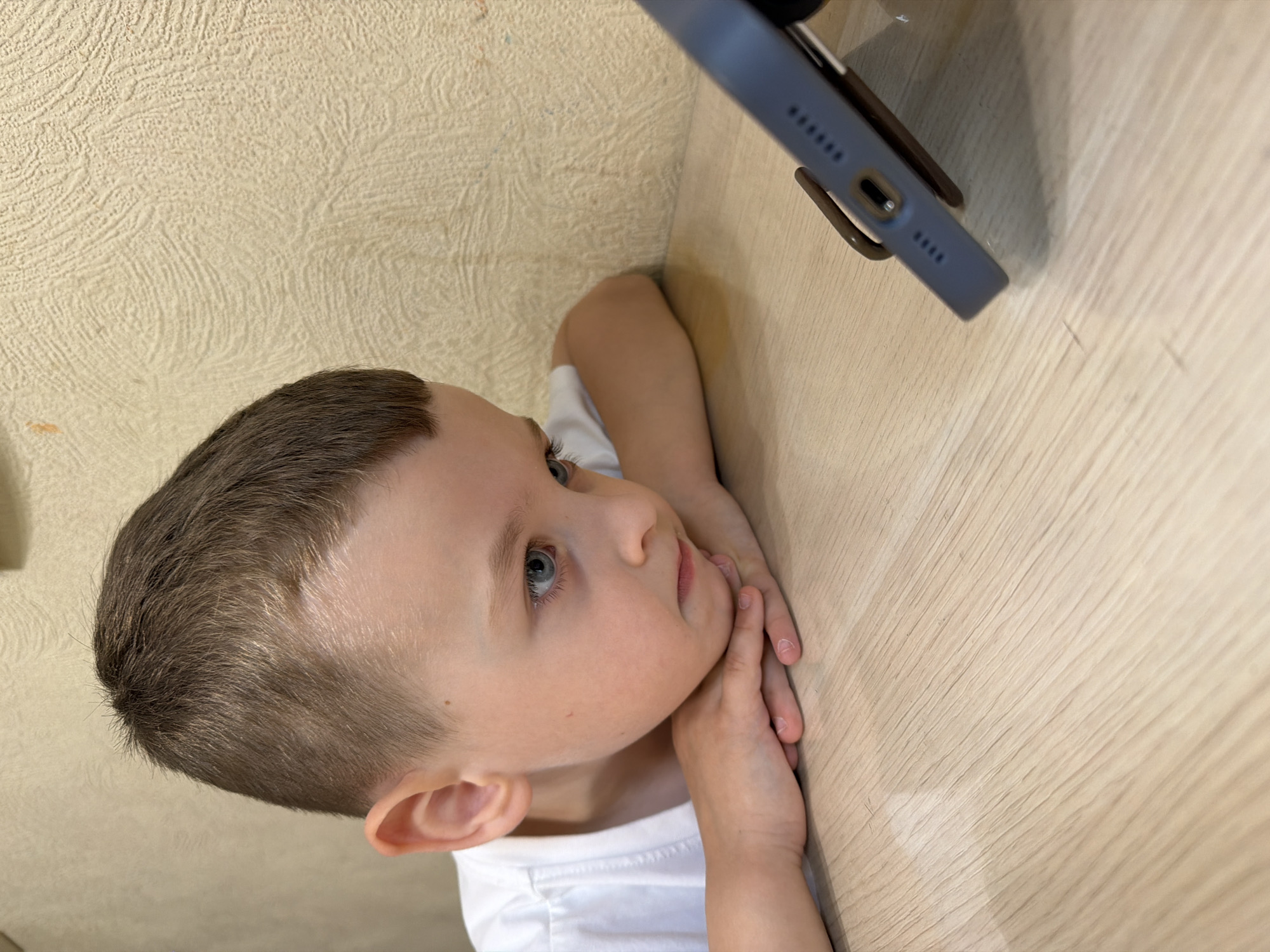A young boy with short brown hair and blue eyes is leaning on his hands, looking intently at a smartphone that is propped up on a stand. The boy is wearing a white t-shirt. He is sitting at a light-colored wooden table, with a textured beige wall in the background. The image appears to be taken indoors, likely in a residential setting, possibly in Khabarovsk, Russia, although there are no specific visual cues to confirm this location. The lighting is soft and diffused.