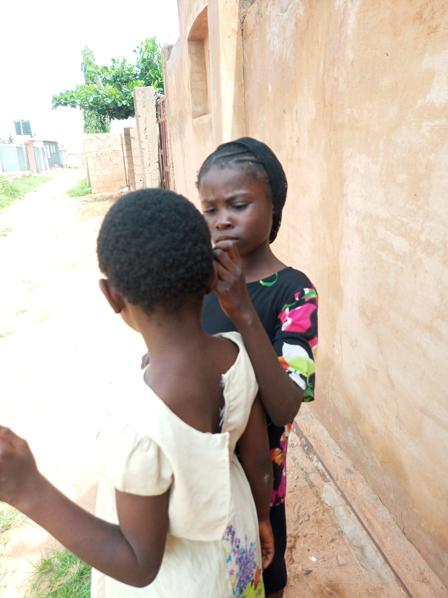 In Tempure, Nigeria, two young girls stand in front of a textured, light-brown wall. The girl on the right wears a black headscarf and a black shirt with a colorful floral pattern, her hand touching her face. She is looking down at the girl on the left, who has short, dark, curly hair and is wearing a light-colored dress with short sleeves. The dress has floral embroidery near the hem. The background shows a dirt path, some green foliage, and a portion of a building with a metal gate. The scene appears to be an outdoor, residential area.