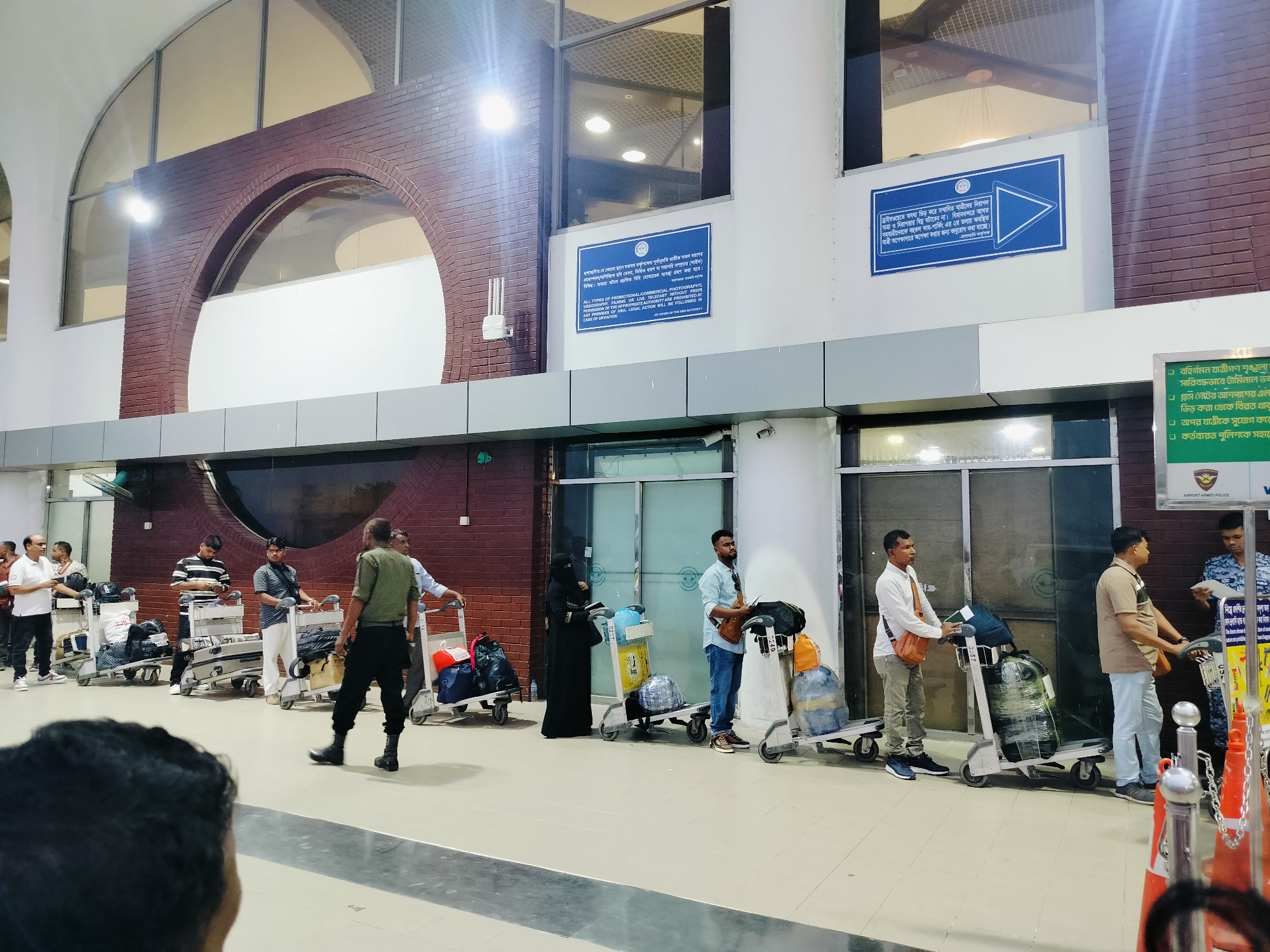 The image depicts a scene inside what appears to be an airport terminal in Dhaka, Bangladesh, specifically at the Hazrat Shahjalal International Airport. The environment features a large, modern interior with high ceilings, arched brickwork features, and reflective floors. Multiple people are present, most of whom are standing with luggage carts.
In the foreground, the back of a person's head is visible, facing away from the camera. Several individuals are positioned behind luggage carts, some appearing to be waiting or in transit. Notable individuals include a man in a military-style uniform walking towards the right side of the frame, and a woman in a black hijab with a luggage cart. Other people are seen interacting with their luggage or standing in lines.
The luggage carts contain various bags, suitcases, and wrapped items. Signs are mounted on the wall, some in Bengali and English, providing information or directions. One sign, partially visible, appears to be a prohibition notice. Another sign displays a blue triangle icon. The overall atmosphere suggests a busy travel hub. The lighting appears to be artificial, with bright ceiling lights illuminating the space.