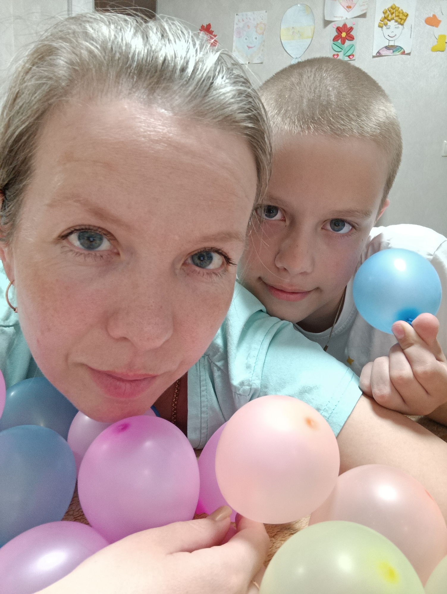 A close-up shot features a woman and a young boy surrounded by light pink, blue, and yellow balloons. The woman has light skin, blue eyes, and light blonde hair pulled back from her face; she is wearing hoop earrings and a light blue shirt. The boy has light skin, blue eyes, and a buzz cut. He wears a white shirt and holds a blue balloon in his hand. Behind them, on a white wall, are several children's drawings. The location is presumed to be an indoor setting in the city of Chaikovsky, Russia.