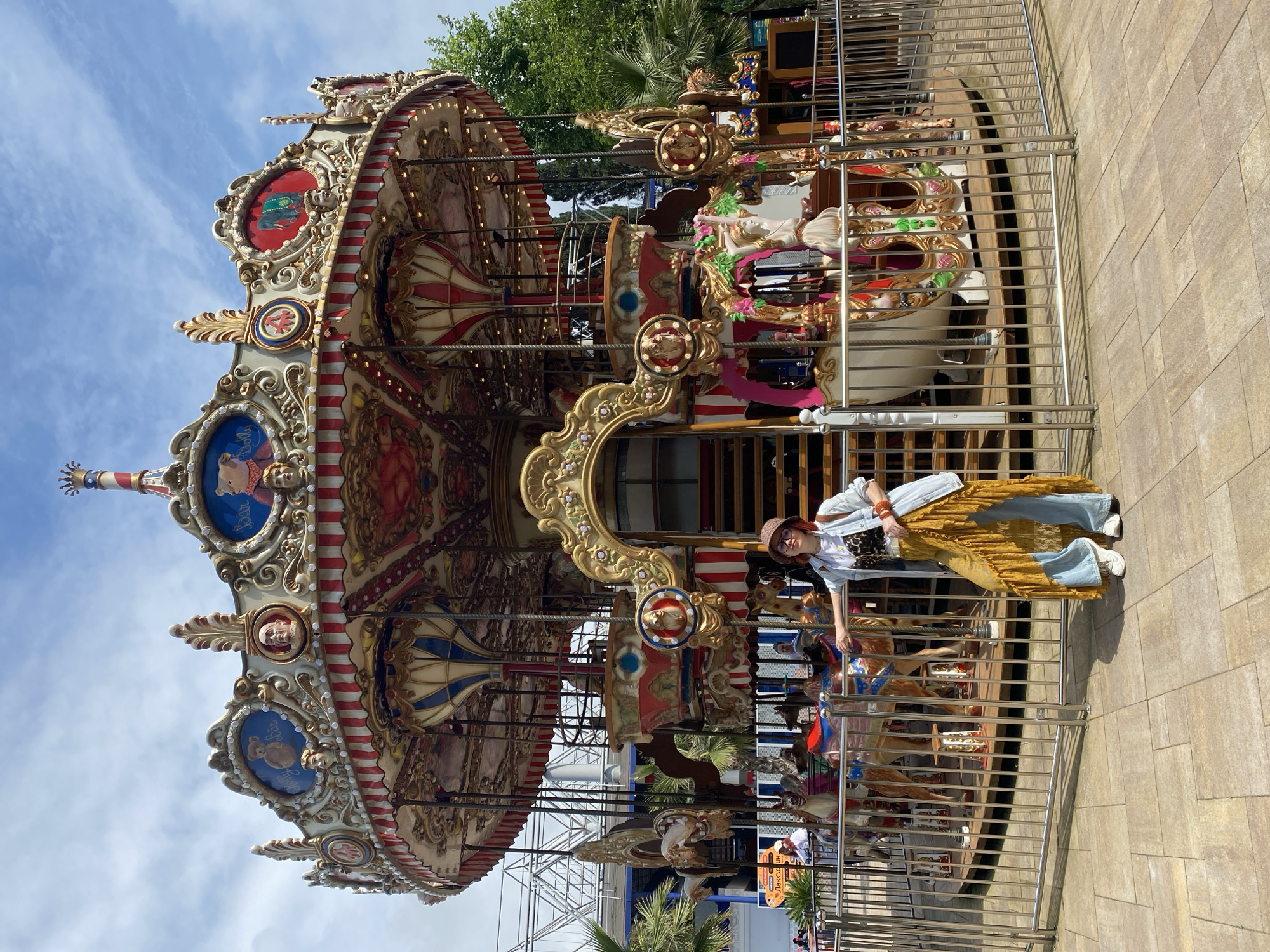 A full shot of a woman standing in front of a carousel. The woman is wearing a hat, glasses, a leopard print top, a denim jacket, a yellow dress, and blue jeans. She is standing with one hand on the railing of the carousel. The carousel is decorated with gold trim and has a variety of horses and other animals on it. The background is a blue sky with some trees. The carousel is located in the city of Sochi, Russia.