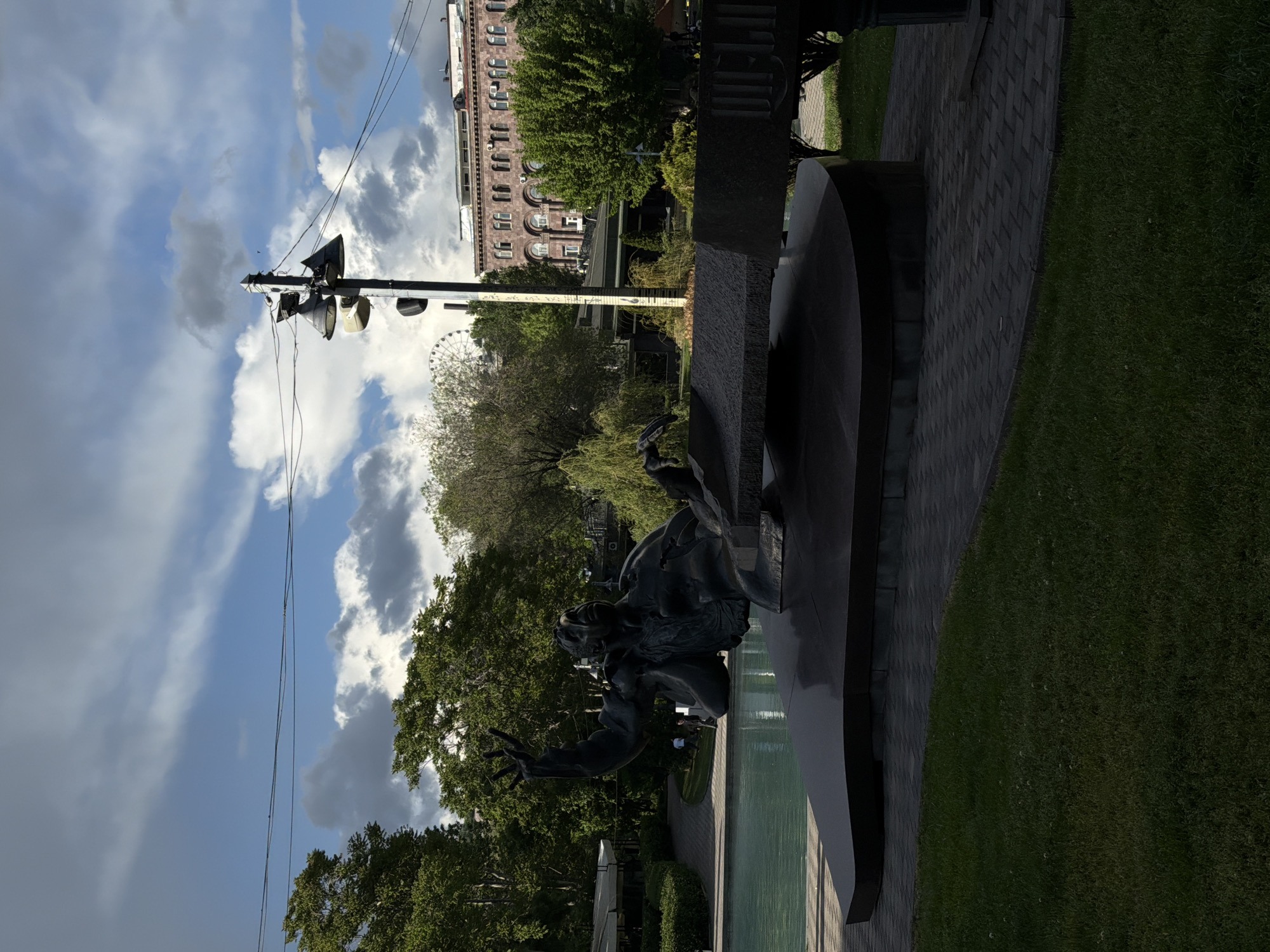 The image shows the statue of "David of Sassoun" in Yerevan, Armenia. A bronze statue depicting the hero David on horseback is in the foreground, with a water feature nearby. Behind the statue, there is a tree-lined park and a Ferris wheel is seen in the background. An overhead light with several lights is visible on the left side. A building with many windows is visible behind the trees. The weather appears sunny with scattered clouds. The image is slightly tilted to the right. There are green grass and brick sidewalks in the scene.