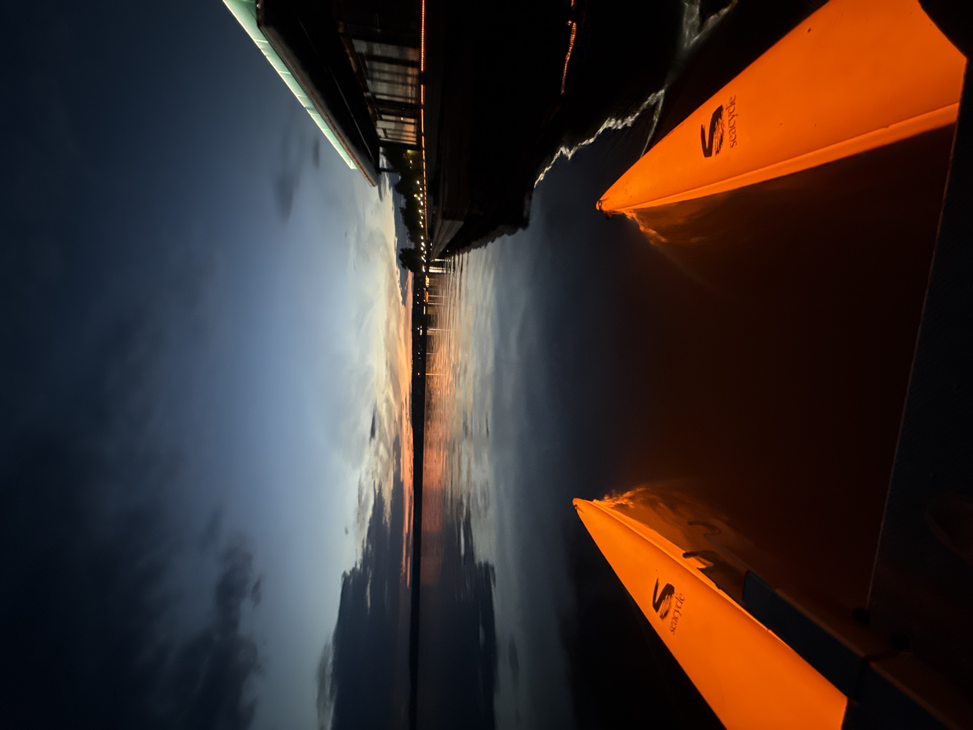 This is a low-angle, first-person perspective shot taken from a kayak or similar watercraft at dusk on Lake Votkinsk, Votkinsk, Russia. Two bright orange kayak outriggers, each with the "Seacycle" logo, are prominently featured in the foreground, extending into the dark water. The lake is calm, reflecting the twilight sky and the lights of a modern building complex on the far shore. The sky displays a gradient of deep blue and dark gray clouds, with a sliver of orange and yellow light on the horizon from the setting sun. The building on the right has large windows and is illuminated from within and by accent lighting along its edge, with a wooden boardwalk in front of it. The overall atmosphere is tranquil and serene, with a hint of futuristic ambiance due to the illuminated kayaks and building. There are no people visible in the image.