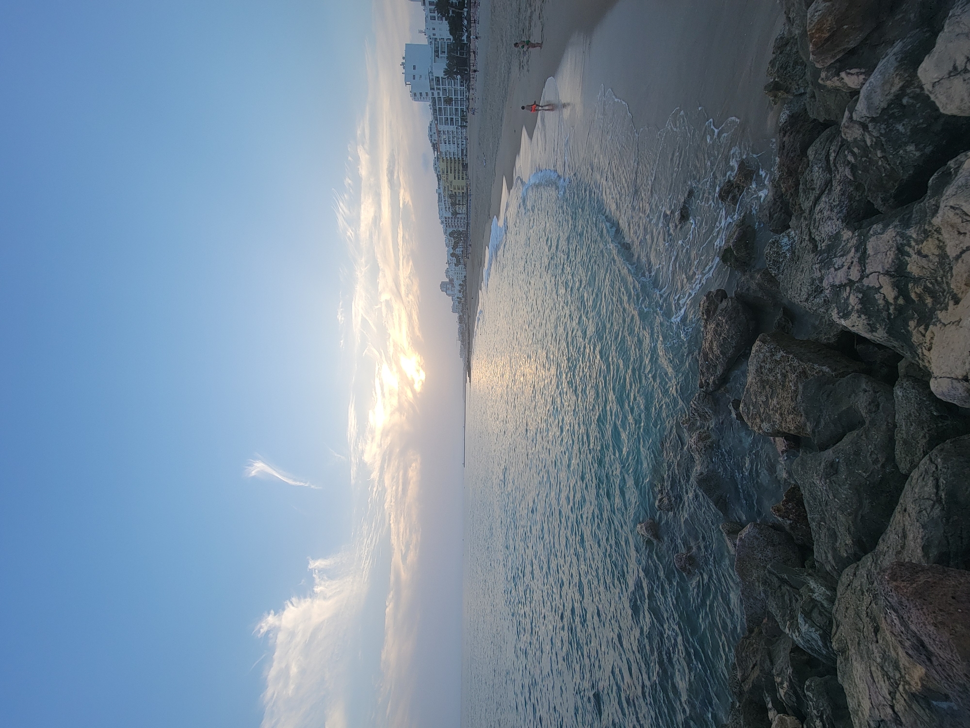 The image depicts a scenic coastal view in Quarteira, Portugal. In the foreground, large, grey rocks line the edge of the water, with the clear, blue ocean stretching out to the horizon. The sea is calm, with gentle waves lapping at the shore. A sandy beach curves along the coastline, dotted with a few people, including a child in an orange bathing suit and an adult in a green swimsuit near the water's edge. Behind the beach, a strip of buildings, primarily white, with balconies and windows, forms the town's skyline. Palm trees are visible interspersed among the buildings. The sky is a soft blue, with wispy white clouds, and the sun is setting, casting a warm glow across the water and illuminating the horizon. The overall atmosphere is serene and peaceful.