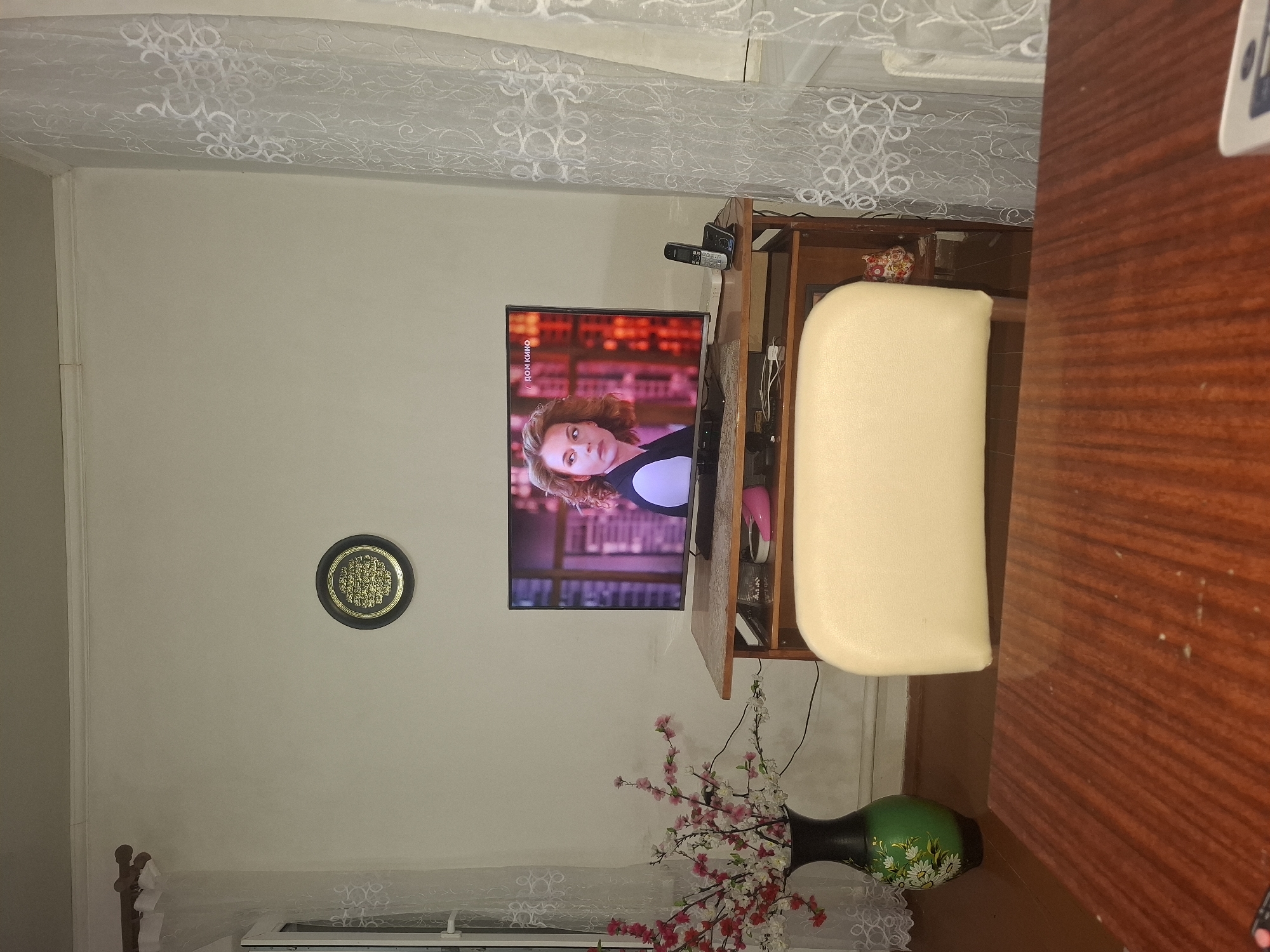 An indoor scene depicts a living room. A television set is mounted on a wooden stand, displaying a woman's portrait. A vase filled with artificial flowers stands to the left of the television, and a cordless phone rests on the stand. A light-colored ottoman is positioned in front of the television stand. A framed Islamic calligraphy hangs on the wall above the television. A wooden table is visible in the foreground. The scene is captured from a slightly elevated perspective. The location is not specified in the image.