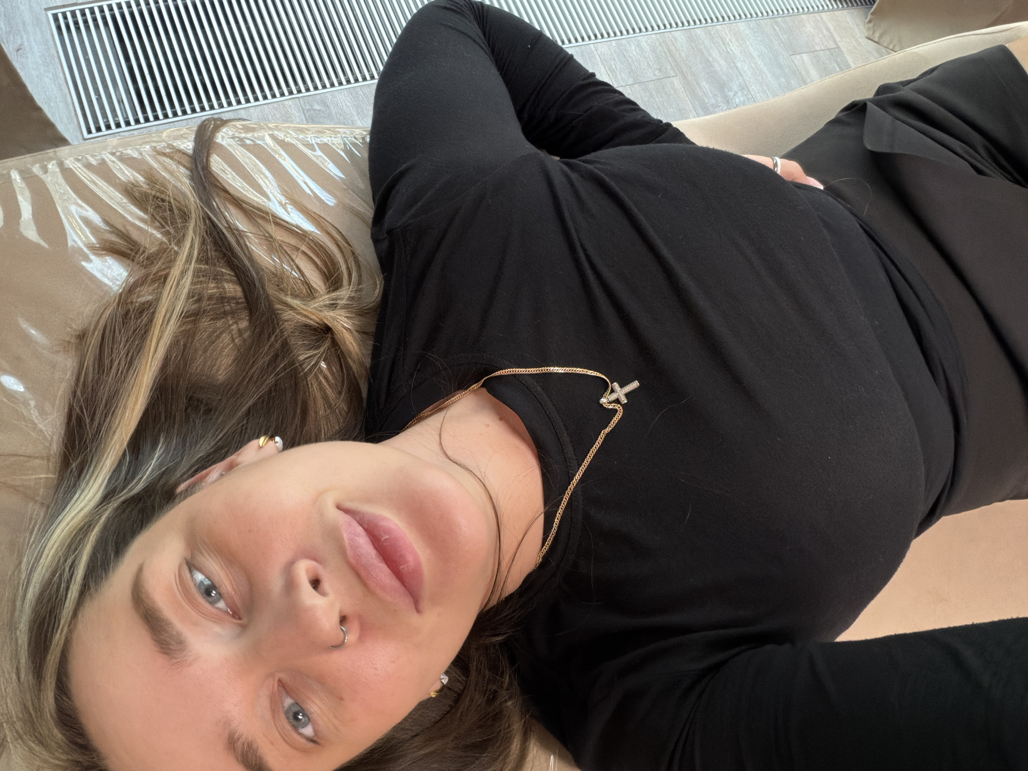 A woman with light hair and blue eyes is lying down, angled towards the camera. She wears a black long-sleeved shirt and a gold necklace with a cross pendant. Her hair is styled with blonde highlights. She has a nose ring and earrings. The setting is indoors, on a beige-colored cushioned surface, possibly a couch. In the background, a white, horizontally-slatted vent or radiator is visible, with light-colored flooring beneath. The image does not show any details that would indicate that the location is Moscow, Russia.
