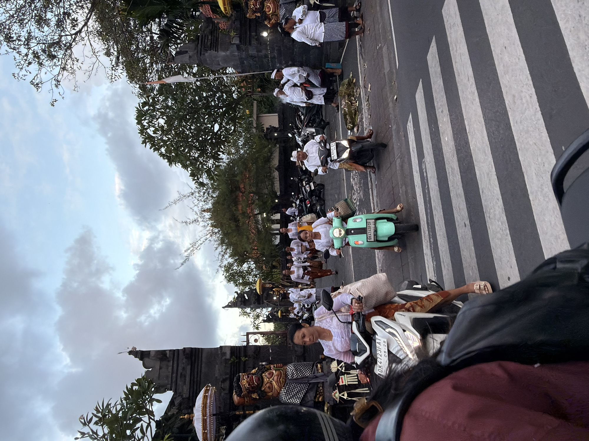 In North Kuta, Indonesia, on a street with a pedestrian crossing, people on motorbikes and walking are seen in front of what appears to be a temple or ceremonial entrance. The sky is overcast, and a cloudy sky suggests it may be late afternoon or early morning. Several people are dressed in traditional Balinese attire, with women wearing white blouses and patterned skirts, and men in white shirts and sarongs with head coverings. One woman on a scooter is wearing a white top and a patterned skirt, while another woman is riding a light green scooter. There are statues and decorative elements visible on the side of the road, including a large, imposing statue dressed in a checkered cloth. The scene depicts a blend of daily life and cultural practices, with motorbikes and a pedestrian crossing indicating a functional urban environment, juxtaposed with religious or ceremonial elements.