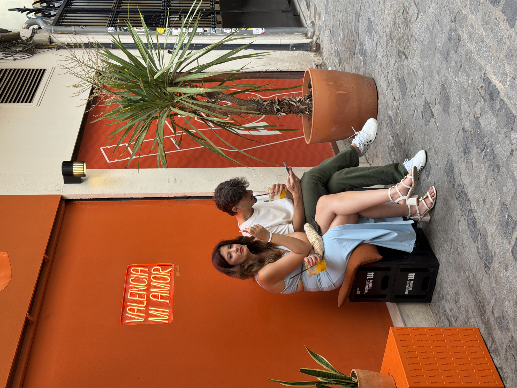Two people are sitting on a bench made of stacked crates outside a building with an orange wall. A neon sign that reads "VALENCIA MI AMOR" is mounted on the wall. The woman on the left is wearing a light blue dress and holding a drink. The man on the right is wearing green pants and a white t-shirt and is looking at his phone. A potted plant is next to them. The ground is paved with cobblestones. The scene appears to be in an urban environment.