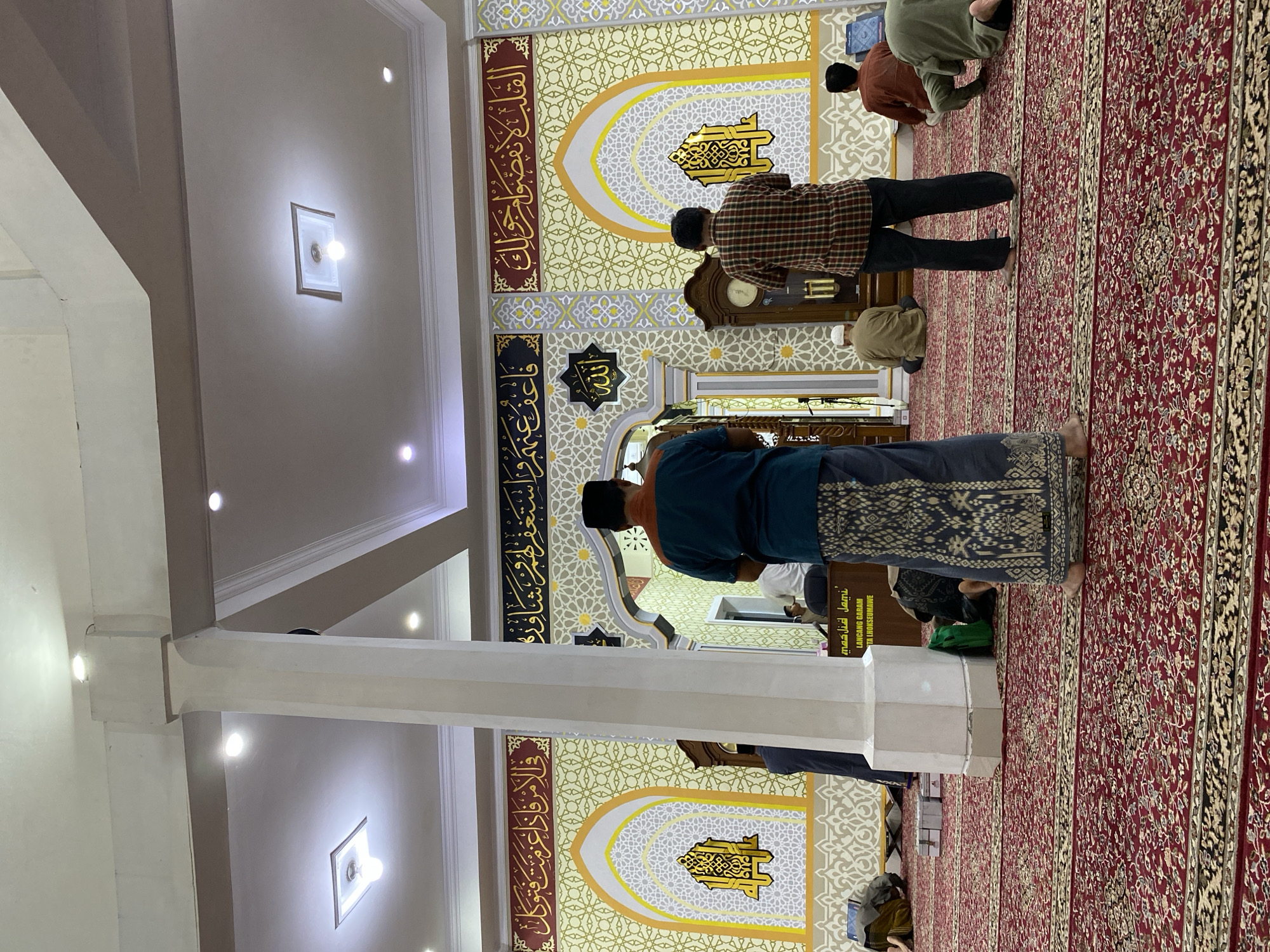 The image depicts the interior of a mosque, likely located in Lhokseumawe, Indonesia, as suggested by signage visible on a wooden structure in the mid-ground. Several men are engaged in prayer, positioned on a red carpet with intricate patterns. A prominent white pillar stands in the foreground, partially obscuring a man facing away from the camera, wearing a blue shirt and a patterned sarong. Behind him, another man in a plaid shirt and dark trousers is also facing away, seemingly in prayer.
The mosque's walls are adorned with decorative Islamic calligraphy and geometric patterns in yellow, gold, and white. Archways with similar decorative motifs are visible on the walls, with one housing a large wooden grandfather clock. In the background, other individuals are seen in various prayer postures, some kneeling or prostrating on the carpet. The ceiling features recessed lighting fixtures. A wooden lectern or counter with visible signage, reading "MASJID JAMI'" and "LANCANG GARAM," is positioned towards the rear of the mosque, near the men in prayer. The overall atmosphere suggests a solemn and devout gathering.