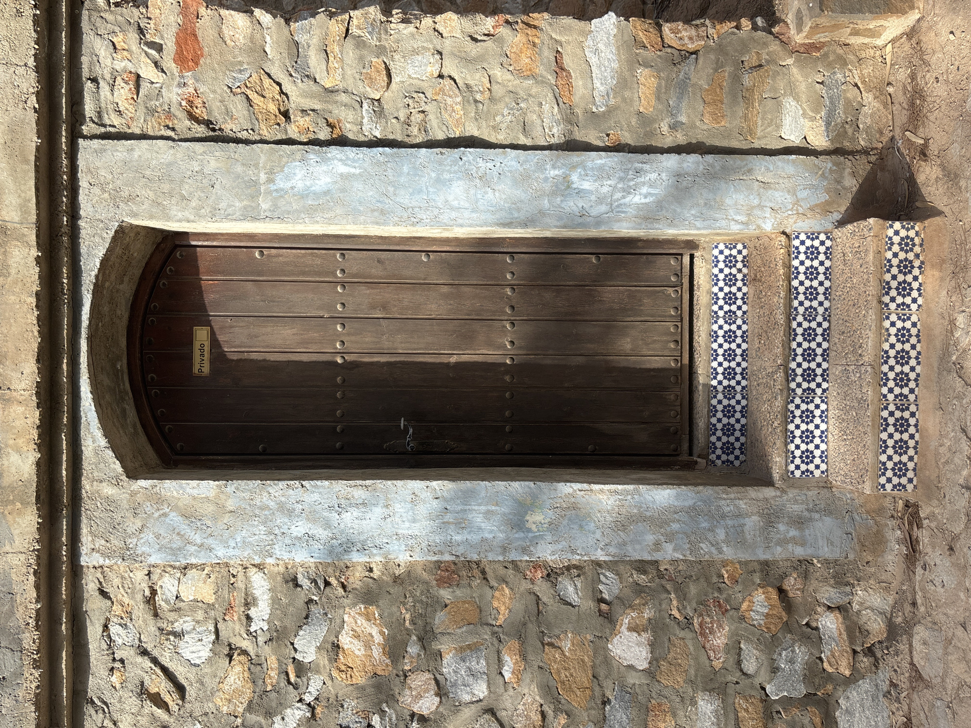 The image shows a rustic wooden door set within a stone wall in Urbanización Cabo Roig, Spain. The door has an arched top and is reinforced with metal studs. A small, rectangular sign that reads "Privado" (Private) is affixed to the upper part of the door. The entrance is accessed by a short set of stairs, each step adorned with blue and white patterned tiles. The wall surrounding the door is constructed from rough-hewn stones of varying sizes and colors, interspersed with mortar. The stonework is partially covered by patches of pale blue and grey plaster. The ground in front of the stairs is made of dirt and small stones. Sunlight casts a shadow across the lower portion of the door and the stairs.