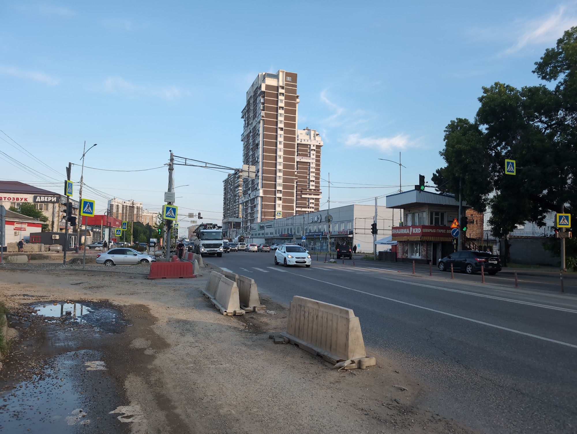 This image captures a street scene in Krasnodar, Russia, likely during daylight hours with a clear blue sky. The foreground shows a construction or repair zone on the side of a road, with a large puddle of muddy water and debris. Concrete barriers and orange traffic barriers are present, indicating ongoing work. Across the road, there is a multi-lane street with traffic, including cars, a truck, and what appears to be a white hatchback.
A prominent feature is a tall, modern, multi-story building with a brown and white facade, set against the sky. Adjacent to it are other buildings, including a low-rise structure with signage in Russian, one of which appears to advertise "SHAURMA." Traffic signals and pedestrian crossing signs are visible. Two individuals are seen on the sidewalk near the shaurma stand, and a person is visible near a dark sedan parked on the side of the road. Trees line the far side of the street, and power lines crisscross the scene. The overall impression is an urban environment undergoing road maintenance.