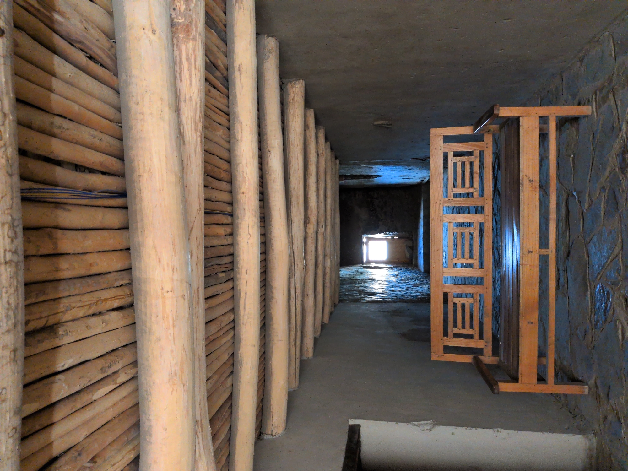 The image shows a wooden bench placed in a hallway with a wooden beamed ceiling. The hallway has rough, textured walls and a stone floor. At the end of the hallway, a doorway is visible, with light coming through it. The bench is positioned centrally in the foreground. The ceiling is constructed from many small, parallel wooden logs, supported by larger, thicker beams. The lighting is dim, except for the bright light emanating from the doorway at the far end. There are no people visible in the scene.