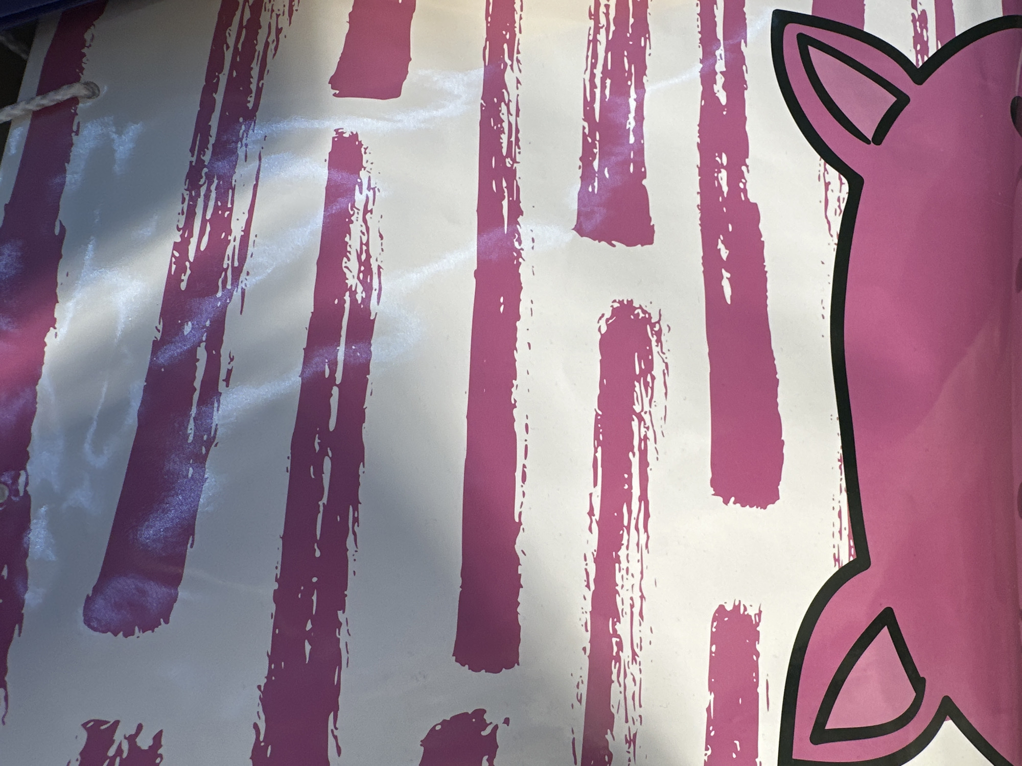 The image is a close-up of a pink and white striped banner, likely a decoration or part of a larger display. The stripes are brushstroke-like, with varying thickness and texture, some appearing solid and others more textured with gaps. Overlaid on the stripes, particularly on the right side of the image, is a large pink shape with a black outline, which appears to be part of a cartoon character's face or head, specifically resembling the ears of a pig or similar animal. A small section of what could be the character's body is also visible. The banner has a rope threaded through a grommet on the top left, suggesting it might be hung. The lighting creates shadows and highlights on the banner, indicating it's illuminated by an external light source, possibly natural light given the dappled shadows. The setting is likely indoors or outdoors in a shaded area, given the light patterns. There is no visible text in the image. The context provided, "Minsk, Belarus," suggests this banner might be for a local event or establishment. The overall impression is festive or decorative, potentially related to a child's party or a themed event.