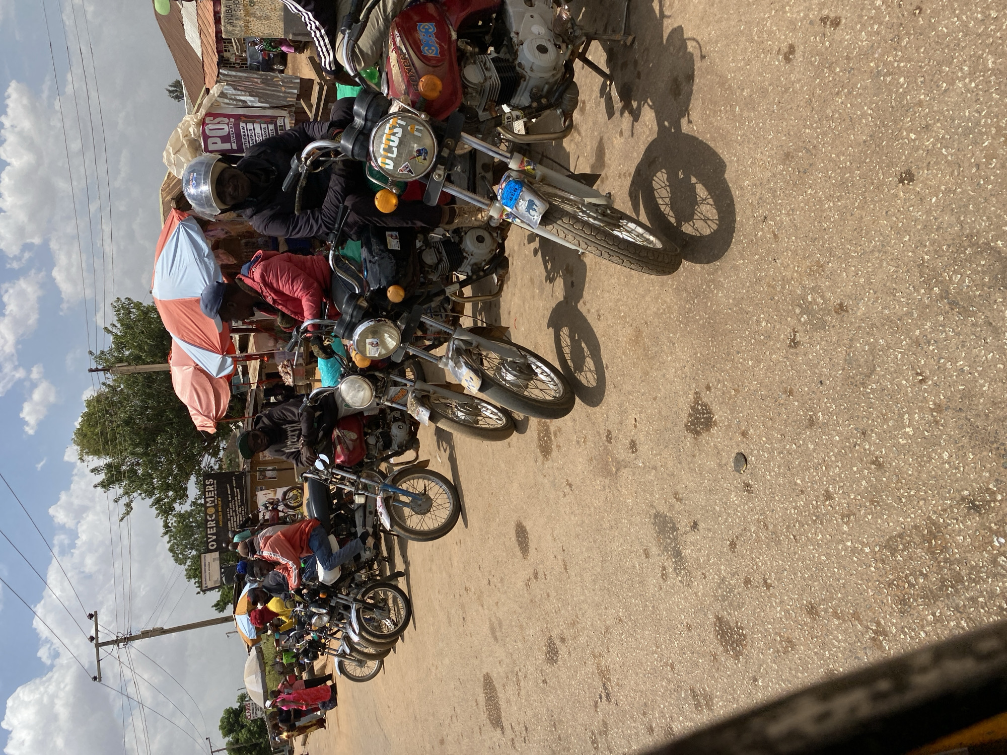 The image captures a street scene in Bukuru, Nigeria, featuring a row of motorcycles parked along the road. Most of the motorcycles have riders sitting on them. The bikes are various models, some appearing older and well-used. A sign in the background reads "OVERCOMERS ANGULDI BRANCH", suggesting a business name and location. Another sign advertises "POS AVAILABLE WITHDRAW TRANSFER DEPOSIT CHARGE BILLS". People are gathered near the motorcycles, some partially obscured. The environment consists of a paved road, a sky with light clouds, buildings and trees in the background. The lighting suggests it is daytime.