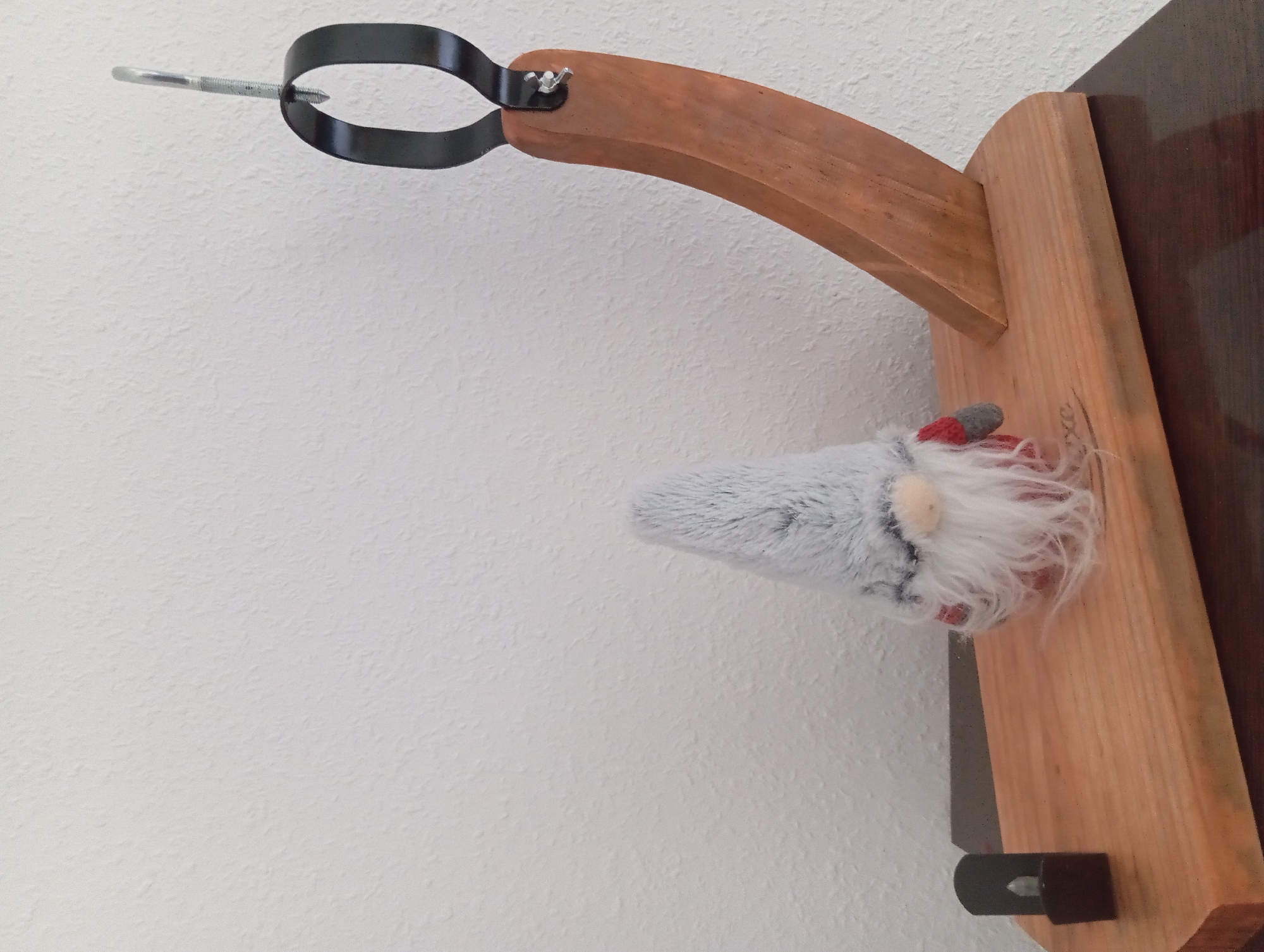 The image depicts a wooden cutting board with a small LED candle on the left and a plush gnome toy on the right. A curved wooden stand with a black metal holder is positioned behind the gnome, suggesting it might be a ham or prosciutto holder. The background is a textured white wall. The scene appears to be set indoors, likely on a countertop or table, given the reflective surface beneath the cutting board. The overall impression is one of domestic decor, possibly with a holiday theme due to the gnome. The specific location in Heitersheim, Germany is not discernible from the image itself, as there are no identifying landmarks or contextual clues.