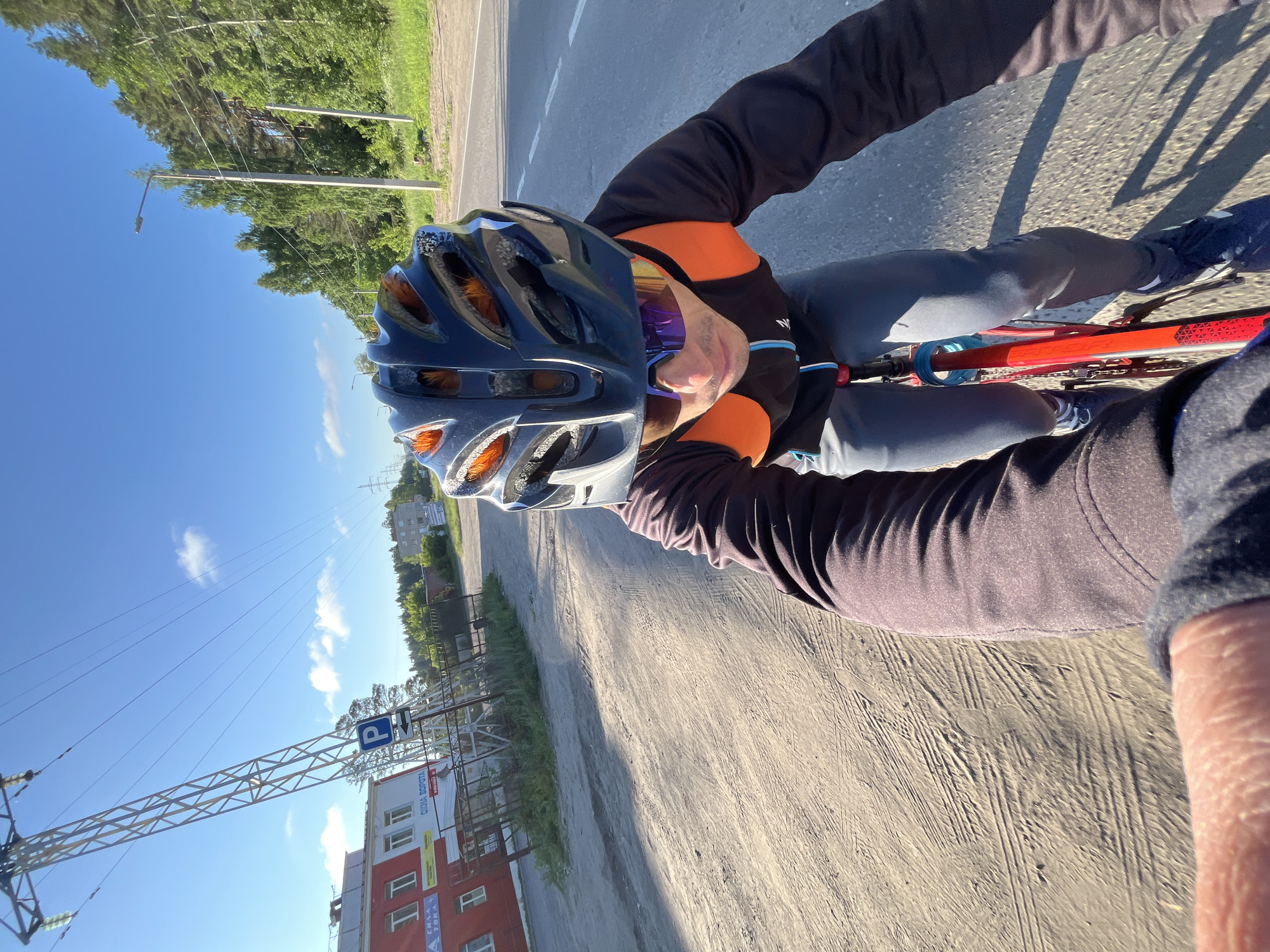 A person wearing a black and orange cycling helmet and sunglasses rides a red bicycle on a dirt road next to a paved road on a sunny day. The person is wearing a black long-sleeved shirt with orange accents and gray pants. In the background, there is a red building with white windows, a parking sign, trees, power lines, and a street light. The scene appears to be on the outskirts of the city of Chaikovsky, Russia, based on the architectural style and vegetation.