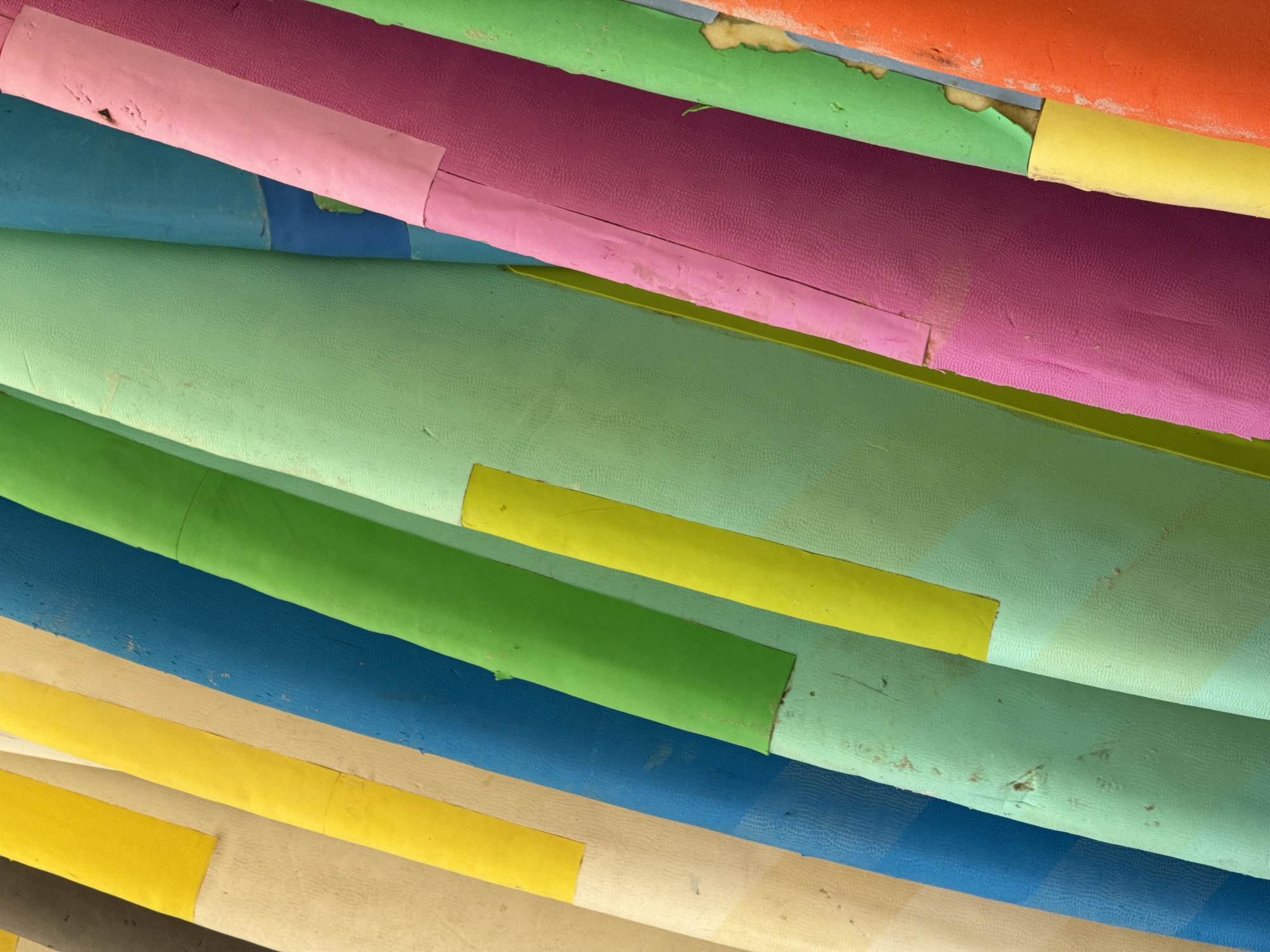 The image is a close-up, diagonal shot of stacked, brightly colored objects, likely rolled materials or surfboards, in Karon, Thailand. The objects are arranged closely together, with their curved surfaces forming vertical lines that recede into the background. The colors include cream, yellow, dark blue, green, light green, pink, purple, and orange. Some of the colored surfaces have yellow or pink rectangular accents taped onto them. The textures appear to be a slightly rough, matte surface, with some scuffing and discoloration visible on certain areas, indicating wear and use. The lighting is even, suggesting an overcast day or a shaded area. There are no people visible in the image.