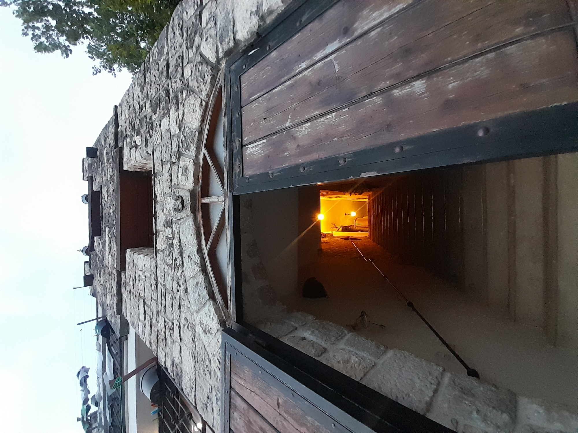 A low-angle shot depicts a stone building with an open wooden door, revealing a tunnel-like interior. The building's exterior is characterized by roughly textured stone walls with small window openings. The wooden door, framed with black metal, is partially ajar, offering a glimpse into the interior. Inside, a long, narrow passage is visible, illuminated by two lights at the far end. A dark object rests near the door opening. The location is Boljevići, Montenegro.