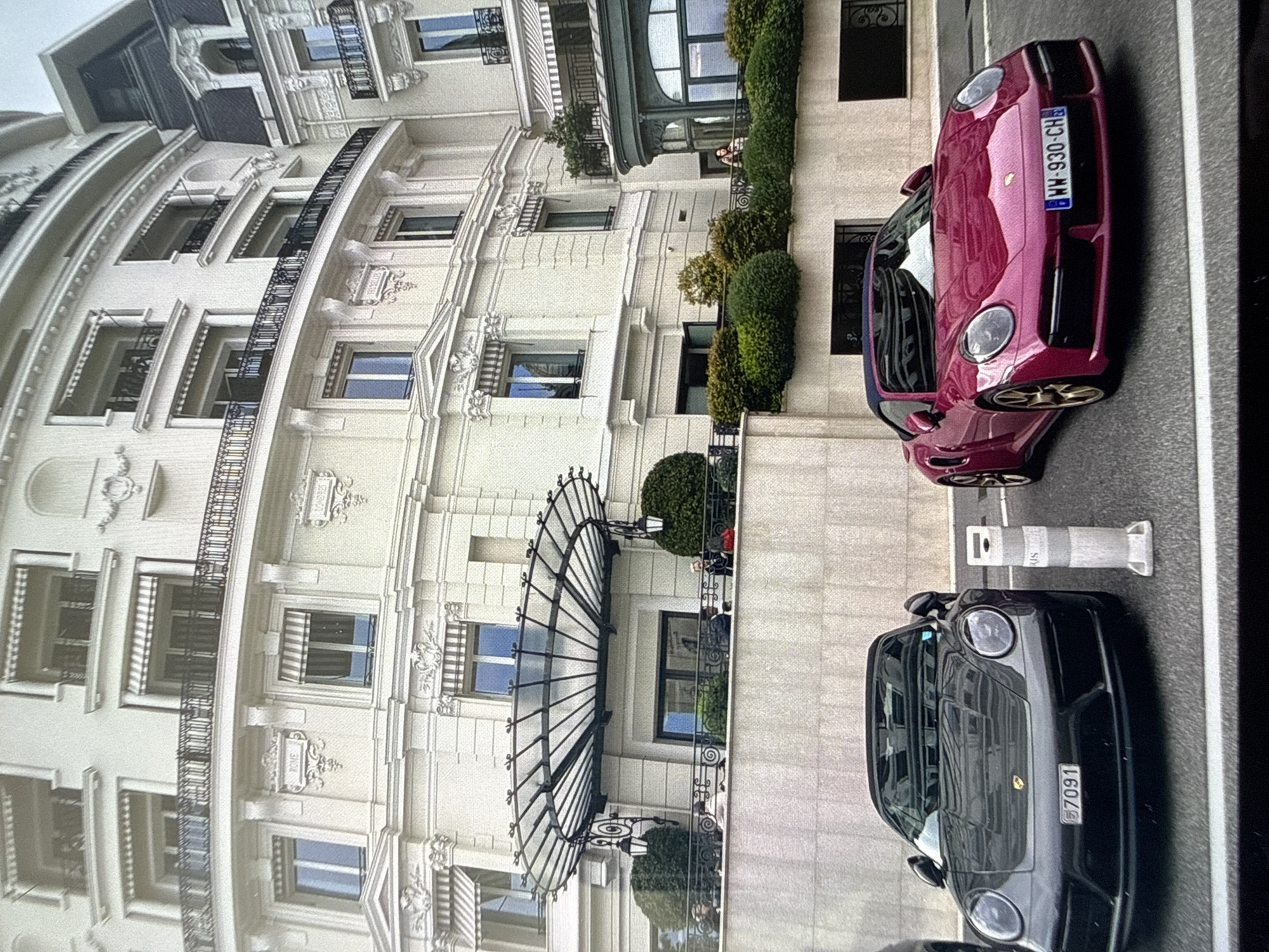 The image shows a European city street scene. Two sports cars, one dark gray and the other burgundy, are parked side-by-side along the street. The cars have license plates visible. In the background is a large, ornate, light-colored building with many windows, balconies, and architectural details. There are green bushes and trees planted in front of the building. The image does not show any clear signs that the photo was taken in the city of Sofia, Bulgaria. The location is uncertain.