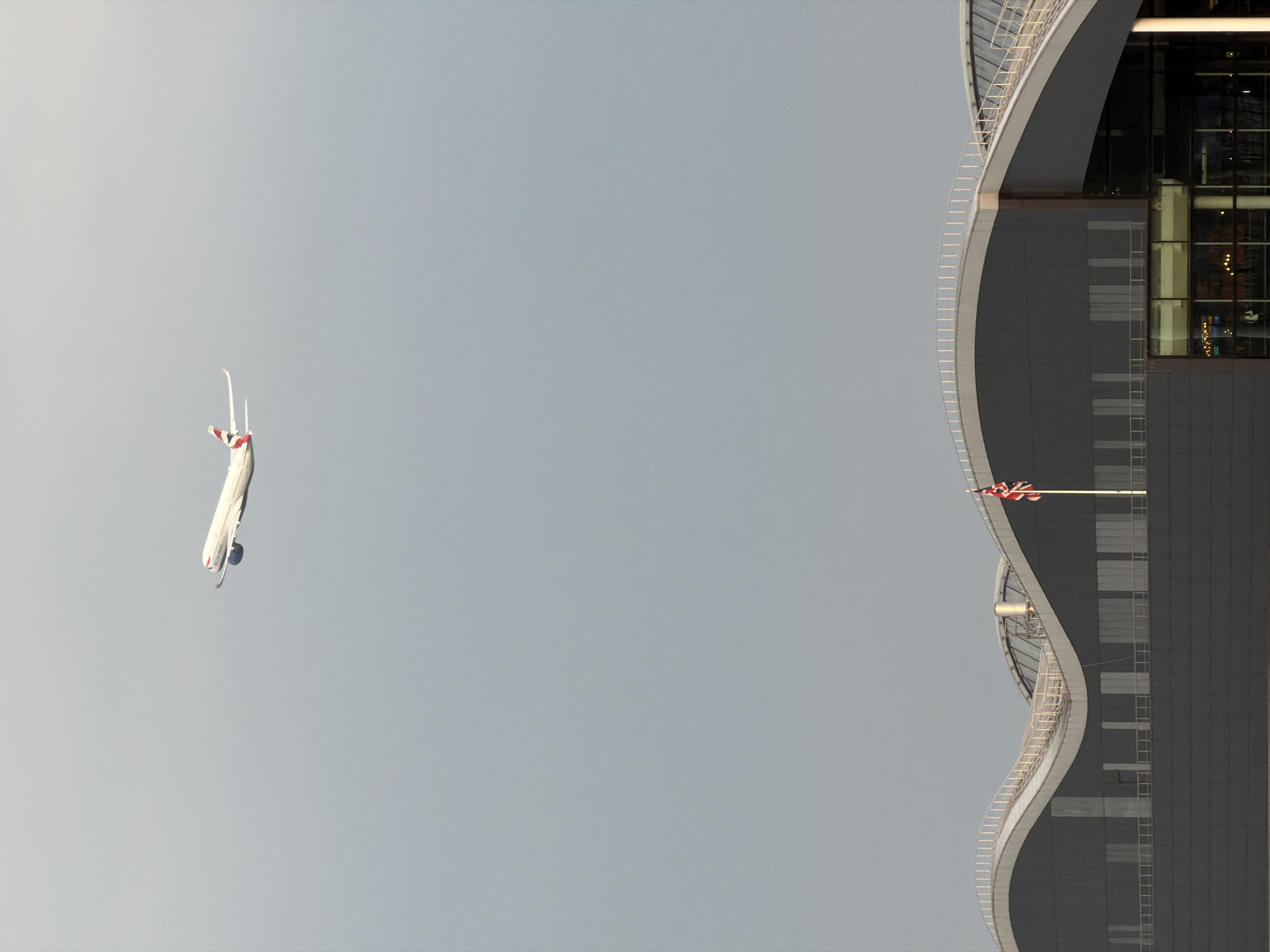 This image, when viewed in its correct vertical orientation, presents a striking composition featuring a large passenger airplane in flight alongside a distinctive modern building under an overcast sky.
On the left side of the frame, a white passenger airplane is captured in mid-flight. The aircraft, identified by the visible text "BRITISH AIRWAYS" on its underside and red markings on its tail and undercarriage, appears to be a large wide-body jet, likely an Airbus A380 given its size. It is flying relatively low, showcasing its underside and a portion of its fuselage against the light grey sky.
To the right, a contemporary building rises with a pronounced, sinuous curve along its facade. The building features dark grey paneling and extensive glass sections, reflecting some ambient light. A white railing or structural element accentuates the graceful curve of its exterior. Attached to the upper part of the building, a flagpole prominently displays the Union Jack, the national flag of the United Kingdom, flapping gently.
The sky is a uniform, soft grey, suggesting an overcast day or hazy conditions, which diffuses the light evenly across the scene. The juxtaposition of the powerful aircraft with the architectural landmark, coupled with the British Airways livery and the Union Jack flag, firmly places the scene in London, United Kingdom, likely near a major airport's flight path. The image captures a dynamic moment, contrasting the movement of the aircraft with the static presence of the urban structure.