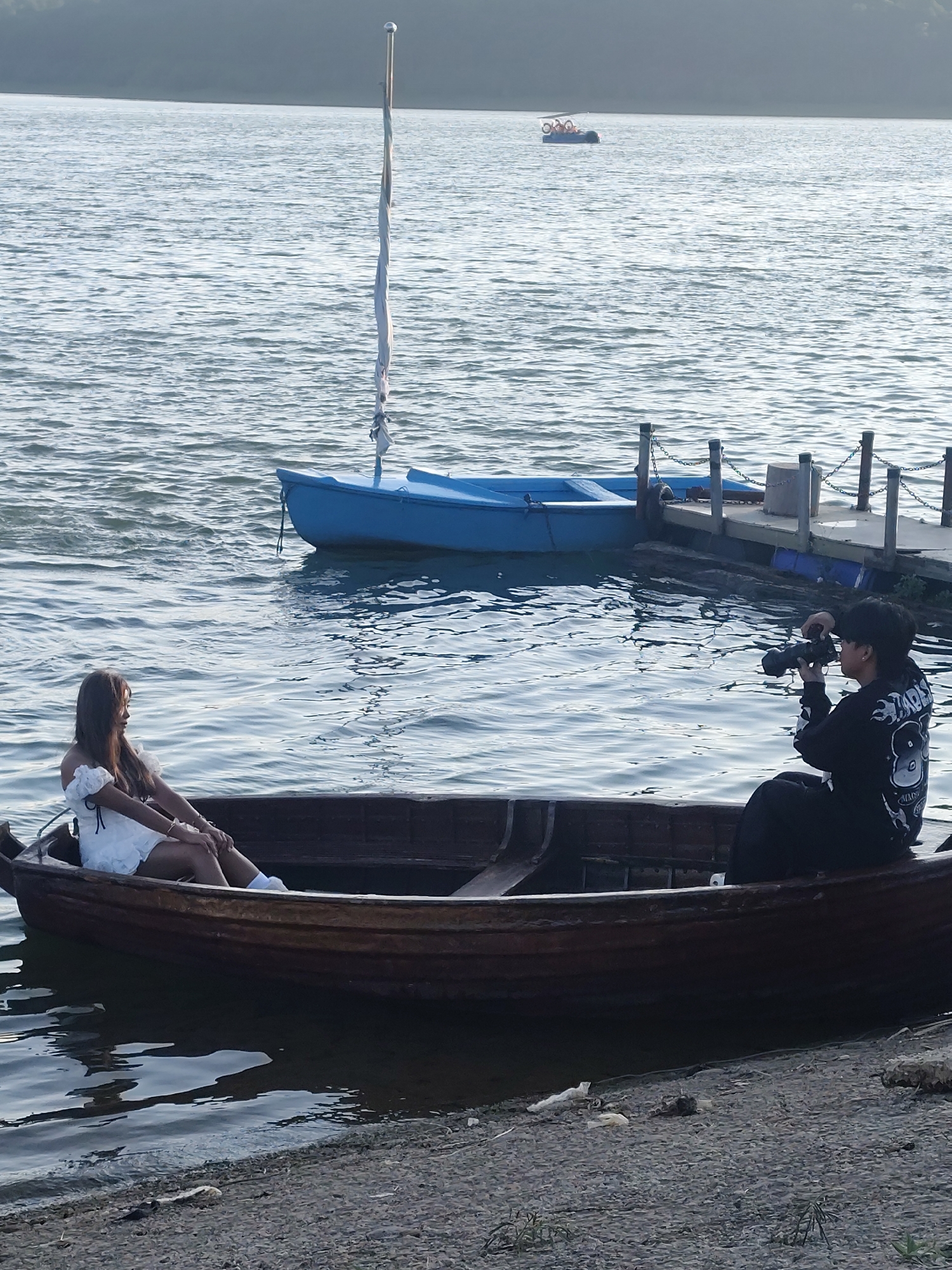 In the city of Unknown, Unknown, on a body of water, a woman in a white dress sits in a wooden boat. She is on the left side of the boat, facing left, with her legs extended. A person in a black hooded sweatshirt and pants is seated in the same boat on the right side, holding a camera and appearing to take a photo of the woman. The boat is near the shore, which is made of gravel. In the background, a blue boat is tied to a wooden pier. Further in the distance, a pedal boat with several occupants is visible on the water. The sky is overcast and the water is rippled.