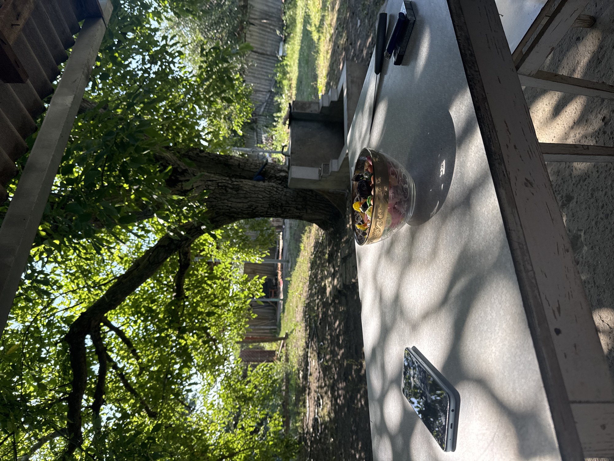 The image depicts an outdoor scene, likely a backyard or patio, in Taganrog, Russia. In the foreground, a light-colored table is visible with a smartphone lying flat on its screen, reflecting sunlight and foliage. To the right of the smartphone, a glass bowl filled with colorful candies or sweets is placed. Next to the bowl are a pack of cigarettes and a lighter. The table casts a shadow with a dappled pattern, indicative of sunlight filtering through tree leaves.
Behind the table, a large tree with dense green foliage dominates the background. The tree's trunk is thick and textured. Beyond the tree, a rustic fence made of wooden planks runs across the yard. The ground appears to be a mixture of grass and dirt, with some circular objects, possibly hoses, lying on the ground. In the middle ground, there is a concrete structure that resembles a well or a water feature with steps leading down to it. The overall environment suggests a warm, sunny day.