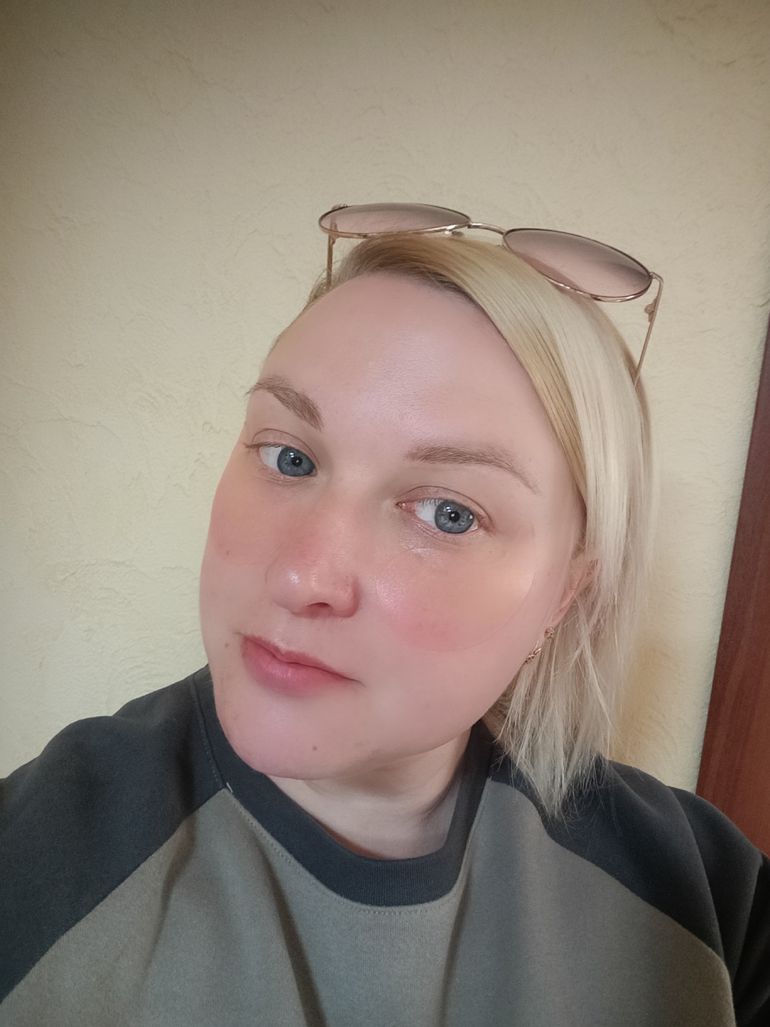 A fair-skinned woman with blond hair is the central subject. She has blue eyes and appears to have eye patches under her eyes. Sunglasses are perched on top of her head. She is wearing a two-toned shirt, with gray sleeves and a beige torso. The background is a light yellow wall. The location is Chaikovsky, Russia. The image appears to be a selfie.