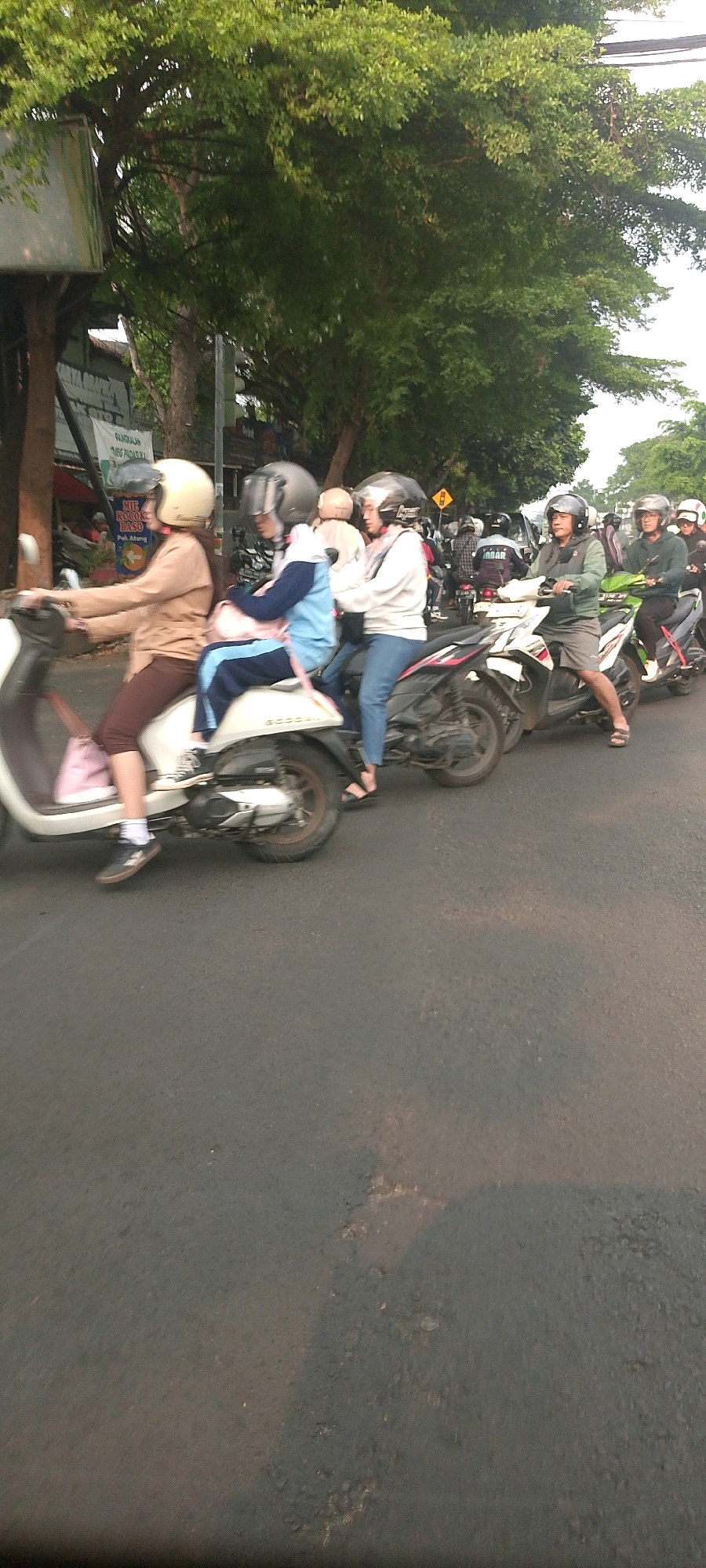 In a city street scene in Bandung City, Indonesia, several individuals are riding on motorcycles. The foreground shows two motorcycles. The first motorcycle, a white scooter, has two riders. The rider in front, a female wearing a light brown long-sleeved shirt and brown pants, has a light yellow helmet. Behind her, a female wearing a blue and white outfit with a pink scarf has a dark gray helmet. The second motorcycle, a dark-colored scooter, also carries two people. The rider in front is a male wearing a green jacket and shorts, with a dark gray helmet. Behind him, a female wearing a white long-sleeved shirt and blue jeans has a dark gray helmet. In the background, other motorcycles and riders are visible, some with helmets and some without. The environment features a tree-lined street with some buildings and signage on the left side. The road surface is asphalt. The lighting suggests it is daytime, possibly late afternoon, with soft sunlight filtering through the trees.