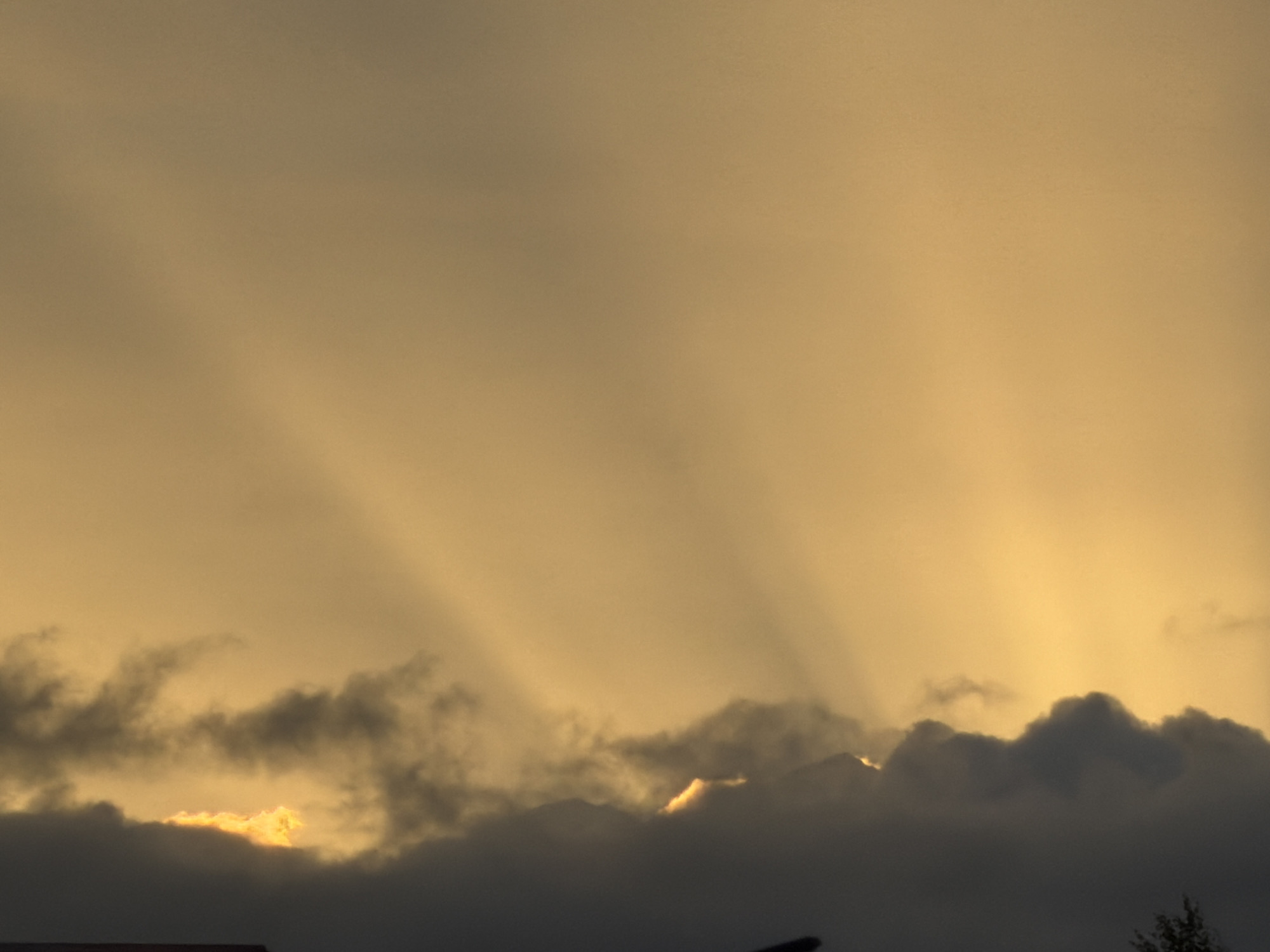 The image shows a dramatic sky over Çolpon-Ata, Kyrgyzstan, likely at sunset or sunrise. Shafts of light, known as crepuscular rays, emanate from behind a layer of dark, ominous clouds that span the lower portion of the frame. The clouds are dense and textured, with some areas appearing wispy and others thick and heavy. A vibrant, fiery orange glow is visible peeking through a gap in the clouds, suggesting the sun is partially obscured. The upper portion of the sky is a hazy, golden-yellow, fading into a lighter tone towards the top. The overall impression is one of atmospheric intensity and natural beauty, characteristic of the region.