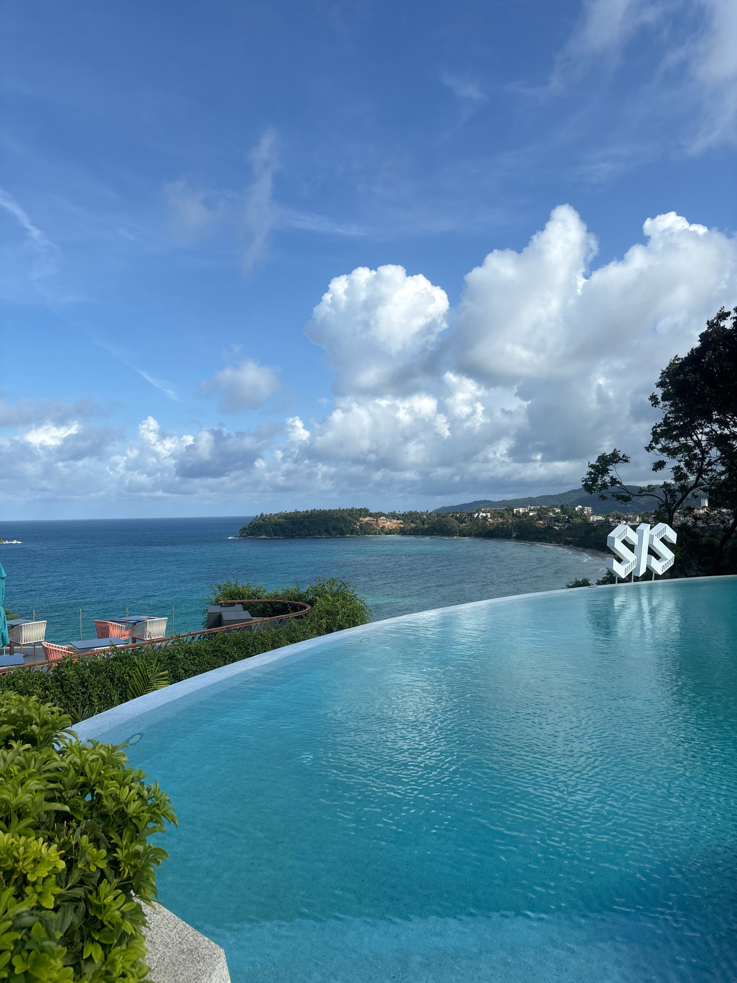An infinity pool overlooks a coastal landscape in Karon, Thailand. The pool's turquoise water reflects the sky, which is a blend of blue with scattered white clouds. In the background, the ocean meets a lush, green coastline dotted with buildings. To the right, a stylized white "SIS" logo stands prominently. Green bushes border the left side of the pool, and in the distance, lounge chairs and tables are visible on a patio area.