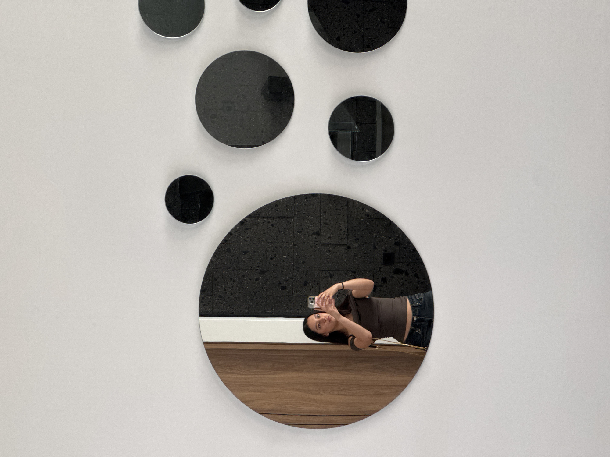 The image depicts a white wall adorned with several round mirrors of varying sizes in an art gallery or installation. A woman is reflected in one of the larger, lower mirrors; she holds a phone in her hands, possibly taking a selfie or picture of the artwork. She is wearing a brown top and jeans. The mirror reflects a wall with a dark speckled pattern above a white baseboard, and below is a wooden floor. The location is Puebla, Mexico.