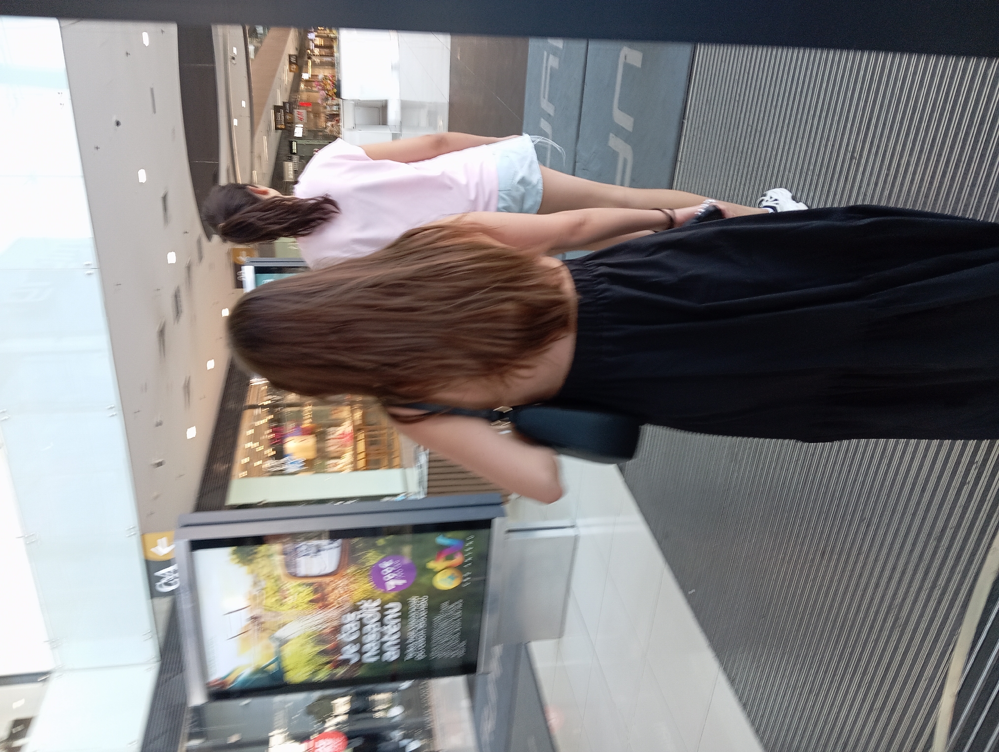 The image shows two young women walking away from the camera in a shopping mall. The woman in the foreground on the left is wearing a black, backless dress and carrying a black purse over her left shoulder. Her long, brown hair is down. The woman on the right is wearing a pink t-shirt and light blue shorts, with her brown hair pulled back in a ponytail. They are walking on a grey, ribbed mat. In the background, there is a large advertisement on a digital display for "otv s antennas" and other shop fronts. The environment appears to be a modern indoor shopping center with bright lighting and polished floors. The "CA" signage visible on a pillar suggests this is likely the OC Mlyny shopping center in Nitra, Slovakia.