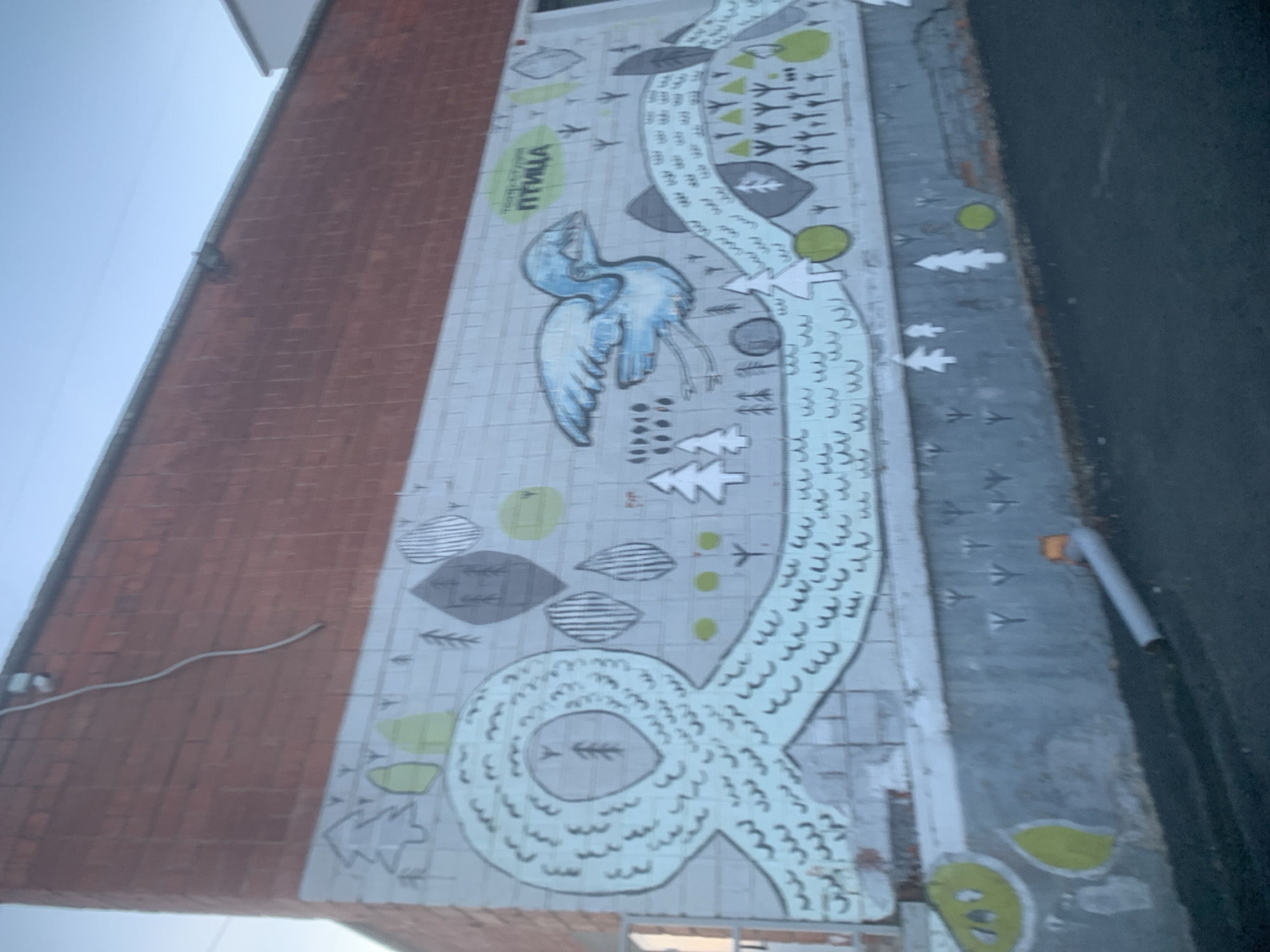 A mural, titled "Bird", adorns the side of a brick building in Izhevsk, Russia. The mural features a blue bird with outstretched wings and stylized leaves and trees in varying shades of gray and green. A winding, river-like shape composed of wave-like lines flows through the mural, containing small tree icons. Additional elements include small silhouettes resembling flying birds. The surrounding environment includes a brick building and a street or sidewalk.