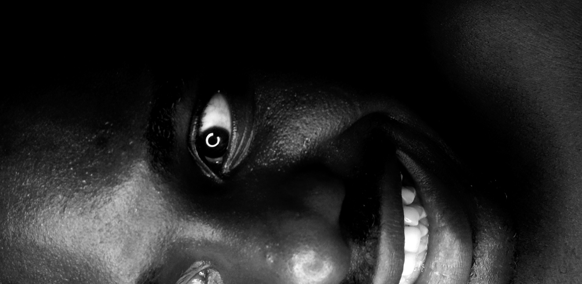 A close-up, black and white image shows a person's face with a distorted perspective, as if viewed from below. The person's eye is visible, with a bright reflection in the pupil. The person's fingers are touching their face, and the white of their teeth is visible. The skin tone is dark. There is no information to indicate this image was taken in Narayi, Nigeria.