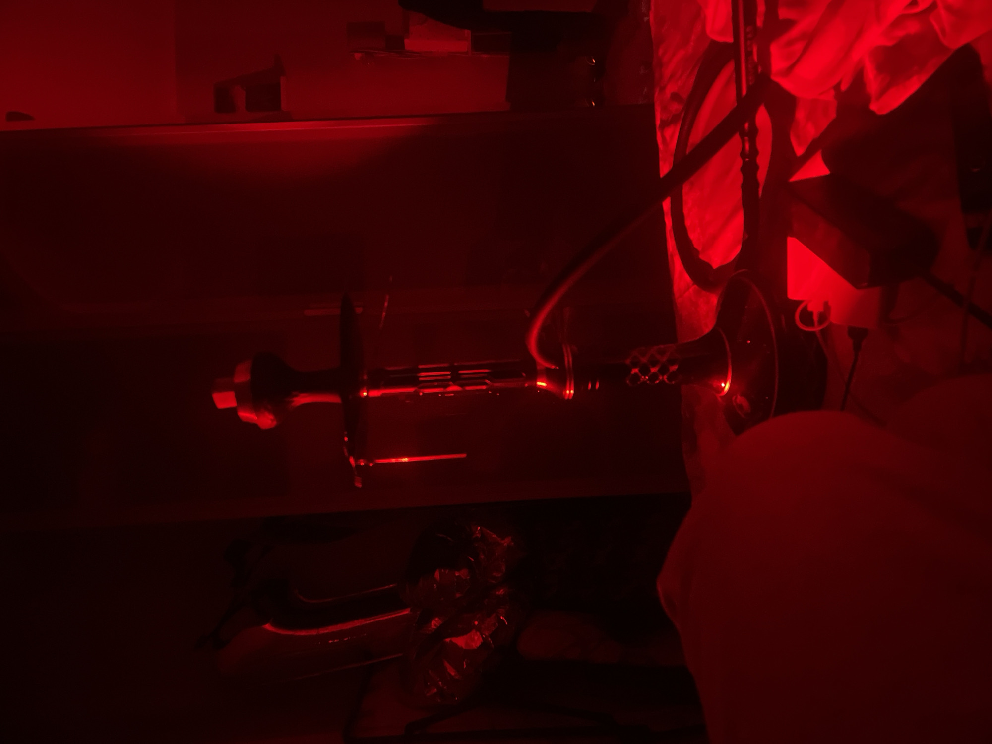 The image depicts an indoor scene bathed entirely in deep red light, creating a low-visibility, atmospheric setting. The primary subject is a metallic hookah (shisha) positioned in the center-left. Its stem is prominently visible, and what appears to be a bowl, possibly covered with foil, sits atop. A hose extends from the hookah towards the right side of the frame. Below the hookah's base, another object, possibly a bundled item or part of a second hookah, is partially obscured and reflects the red light. To the far right, a portion of a person's leg or thigh is visible, clad in light-colored fabric that appears red under the lighting. The background consists of indistinct shapes and shadows, suggesting furniture or other room elements. No specific activity is clearly depicted, but the presence of the hookah implies preparation for or active use of the device. There are no visible faces or identifiable individuals. The lighting strongly suggests an evening or nighttime setting, or a room specifically designed with red ambient lights. No visible text or discernible location cues are present within the image.