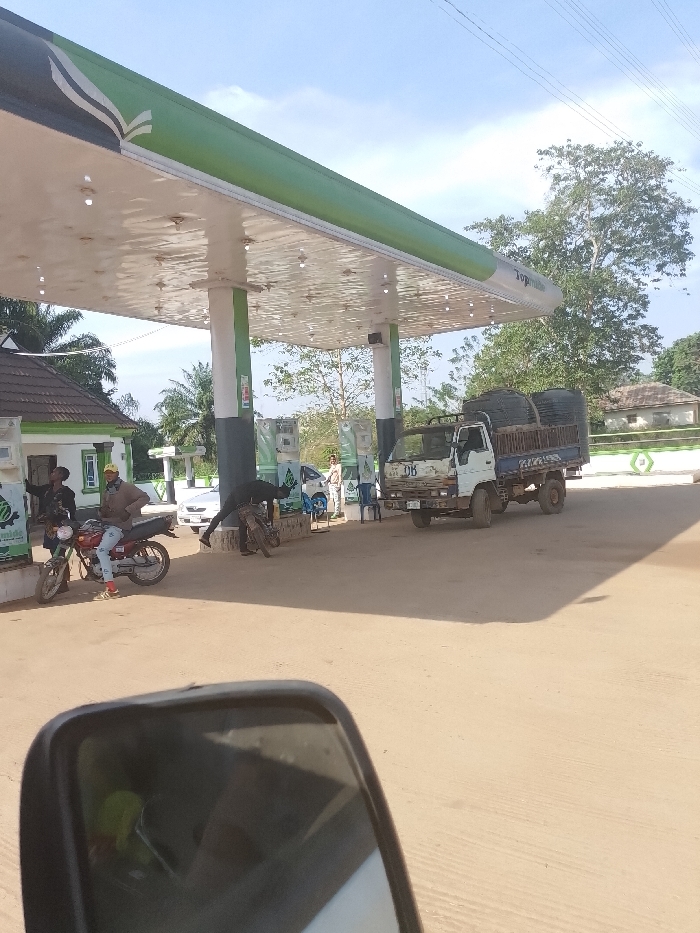 A daylight scene at a gas station in Ayegbaju, Nigeria, is depicted. The primary structure is a large, green and white canopy with multiple white support pillars and an illuminated underside. The gas station's name, "Topman," is visible on the canopy's edge. To the left, a small, white building with a brown tiled roof and green trim, likely an office or convenience store, stands adjacent to the canopy.
The forecourt is a light-colored, open area under the canopy. Fuel pumps, green and white, are positioned beneath the canopy. Several vehicles and individuals are present. On the left, a red motorcycle is parked, with a person wearing a light jacket and yellow cap seated on it. Another individual in dark clothing stands nearby. A third person, also in dark attire, is bent over a darker motorcycle located closer to the fuel pumps.
Further to the right, a weathered white and blue flatbed truck, marked "DB" on its side, is parked under the canopy. Its bed contains several large, dark cylindrical objects. A white car is partially visible behind the fuel pumps, to the left of the truck. Additional individuals are visible in the background near the fuel pumps and the white car. The sky is clear with sparse clouds. In the far background, trees and a building with a brown roof are visible. The lower foreground of the image is partially obscured by a vehicle's side-view mirror.