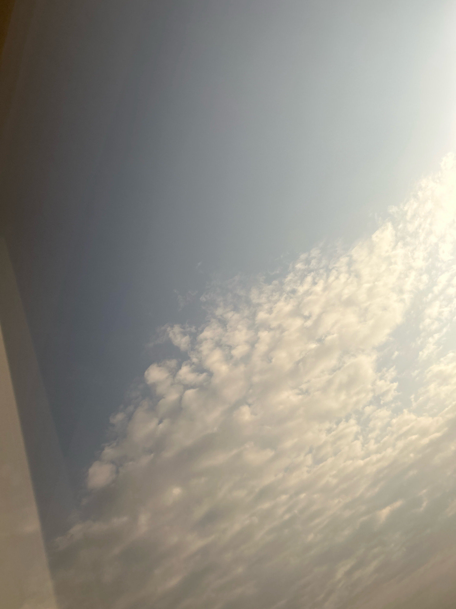 The image depicts a sky filled with clouds, likely taken from an elevated perspective such as an airplane window. A shadowed area, possibly a wing or a part of the aircraft, occupies the left portion of the frame. The right side of the image is dominated by a dense layer of white and grey clouds, illuminated by sunlight that creates a bright, hazy effect. The clouds are textured and varied in form, suggesting a dynamic weather system. The overall scene is one of an aerial view looking up at the sky. There are no visible landmarks or elements that can be used to identify the specific location within Grozny, Russia, or any other city.