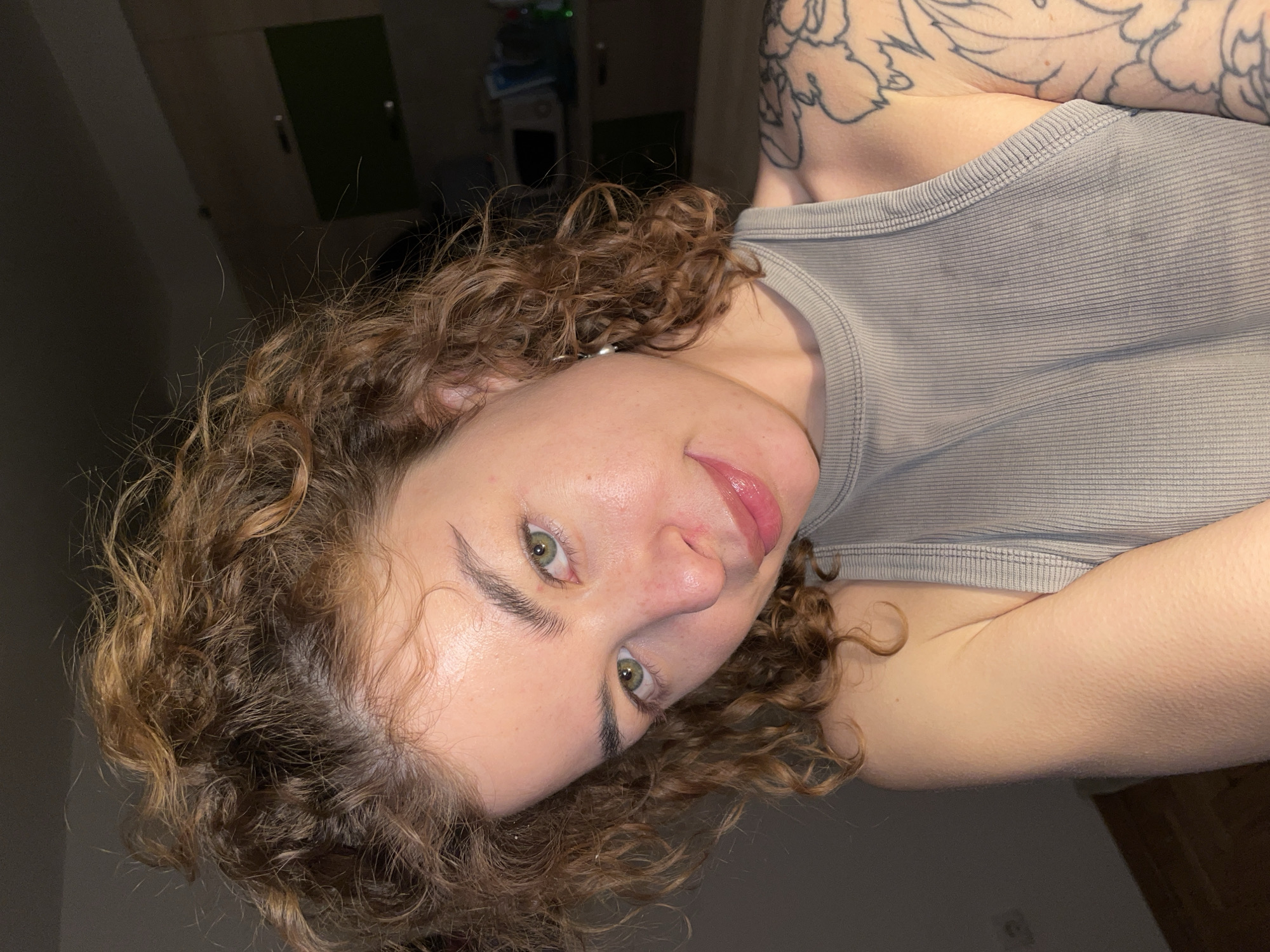 A close-up, angled shot depicts a fair-skinned young woman with light brown, curly hair and green eyes. She is wearing a light grey, ribbed tank top. A tattoo is visible on her upper chest/shoulder area. The background is blurry and appears to be an indoor setting with dark cabinets. The location in Boreti, Montenegro is not discernible from this image.