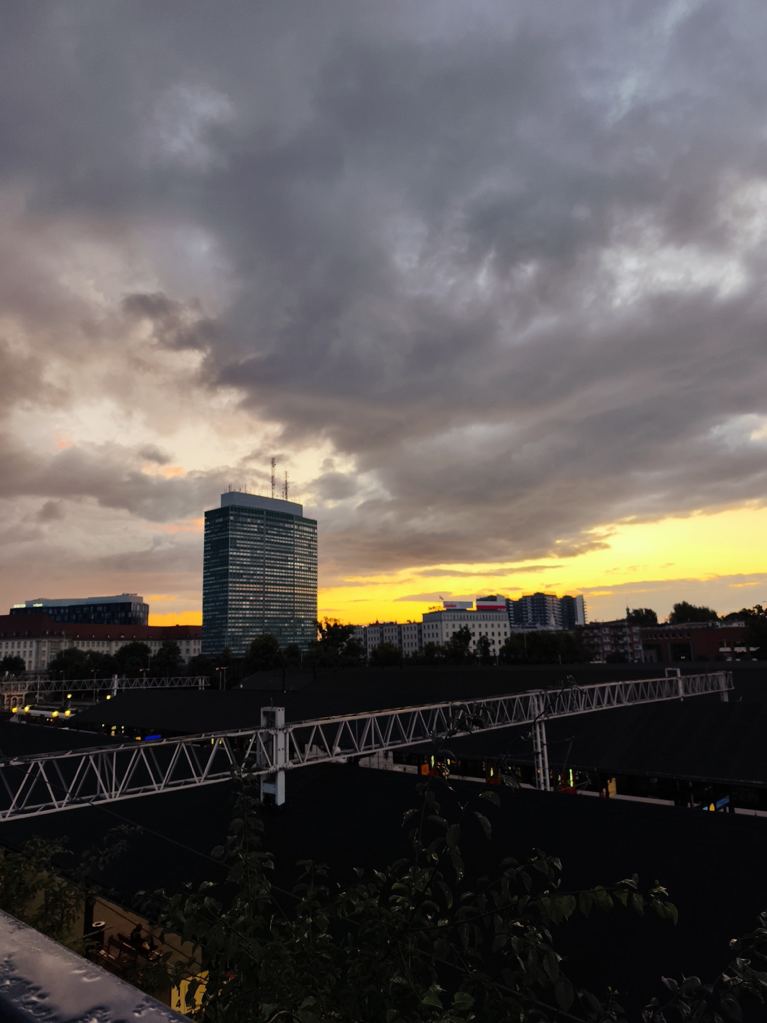 The image captures a dramatic sunset over the city of Gdańsk, Poland. Dark, heavy clouds dominate the sky, tinged with orange and yellow hues from the setting sun. In the mid-ground, a prominent, tall, modern building with a grid-like facade stands out against the colorful sky. To its left and right, other buildings of varying heights and architectural styles are visible. In the foreground, a dark, flat expanse stretches across the lower half of the image, likely a rooftop or open area. A white, industrial-looking structure, possibly a truss or scaffolding, runs horizontally across this dark expanse, its geometric design contrasting with the organic shapes of the clouds and trees. Some greenery is visible in the far foreground, obscuring the lower part of the scene. The overall impression is one of an urban landscape under a powerful and moody sky.