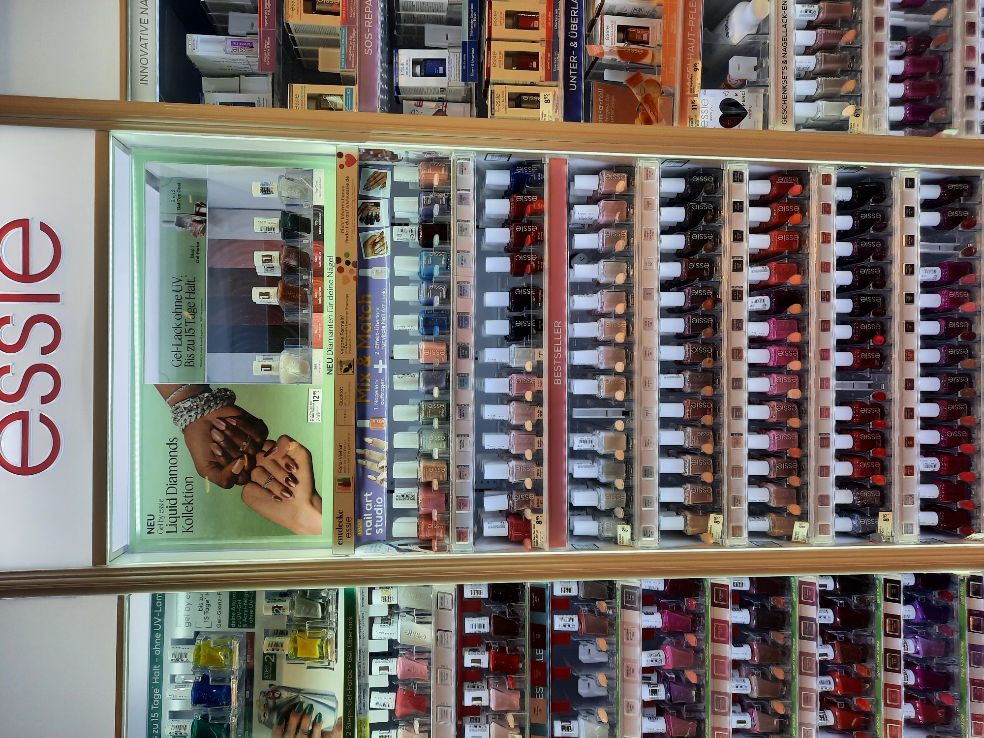 The image shows a retail display of Essie nail polish in a store. The display is brightly lit and features multiple shelves filled with various shades of nail polish bottles. The Essie logo is prominently displayed at the top of the unit.
The display is divided into two main sections. The left section showcases a selection of nail polish bottles organized by color and potentially by product line. There are also promotional materials and signage indicating product features. The right section displays more nail polish, along with some larger treatment or care products.
In the background, other retail displays and store fixtures are visible, suggesting a beauty or cosmetics section of a larger store. The overall environment appears to be a well-organized and brightly lit retail space.
The location is stated as being in the city of Rosenheim, Germany.